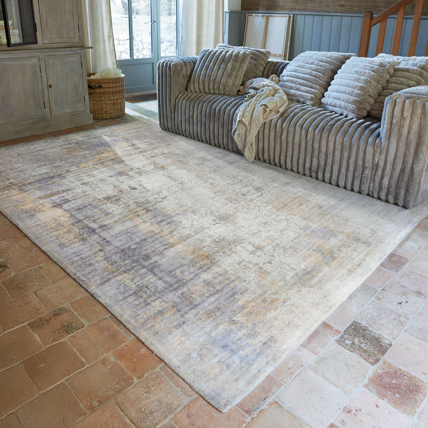 Teppich Qalheera Großer Teppich in Beige- und Grautönen auf Ziegelboden in gemütlichem Wohnzimmer mit grauem Sofa