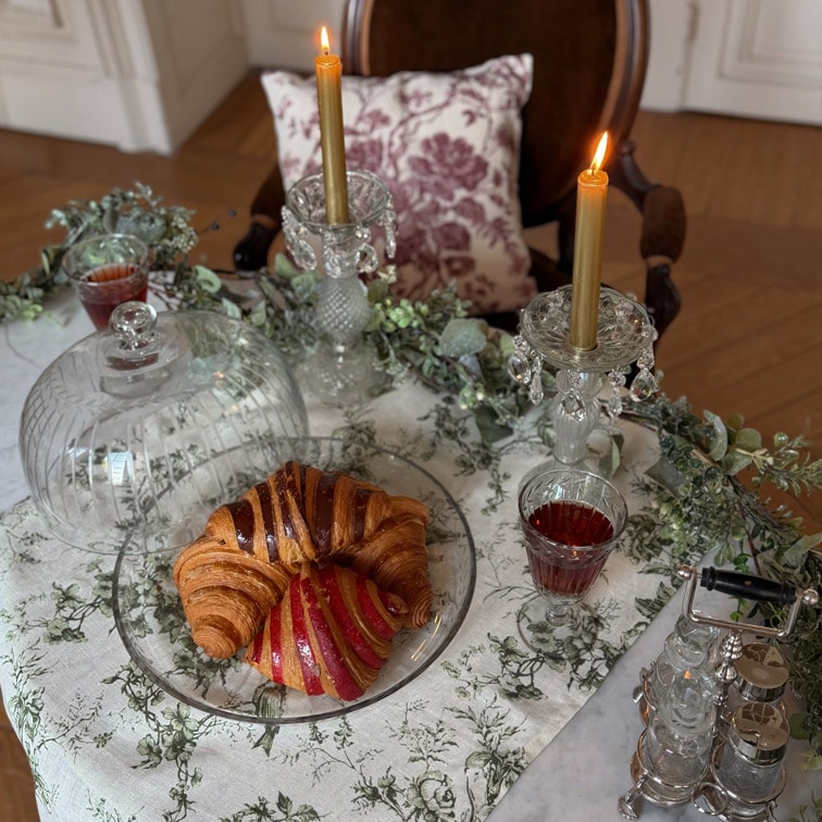 Glassteller mit Croissant und Gebäck auf Tisch mit weißem Tischtuch, zwei goldene Kerzen in Kristallkerzenhaltern