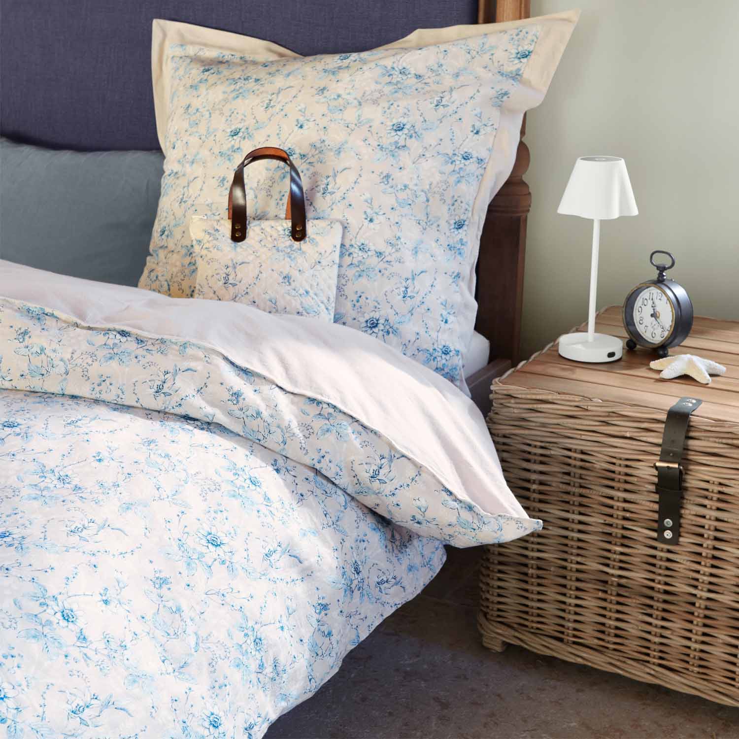 Helle Baumwollbettwäsche mit blauem Blumenmuster auf Bett neben einem Nachttisch aus Korbgeflecht