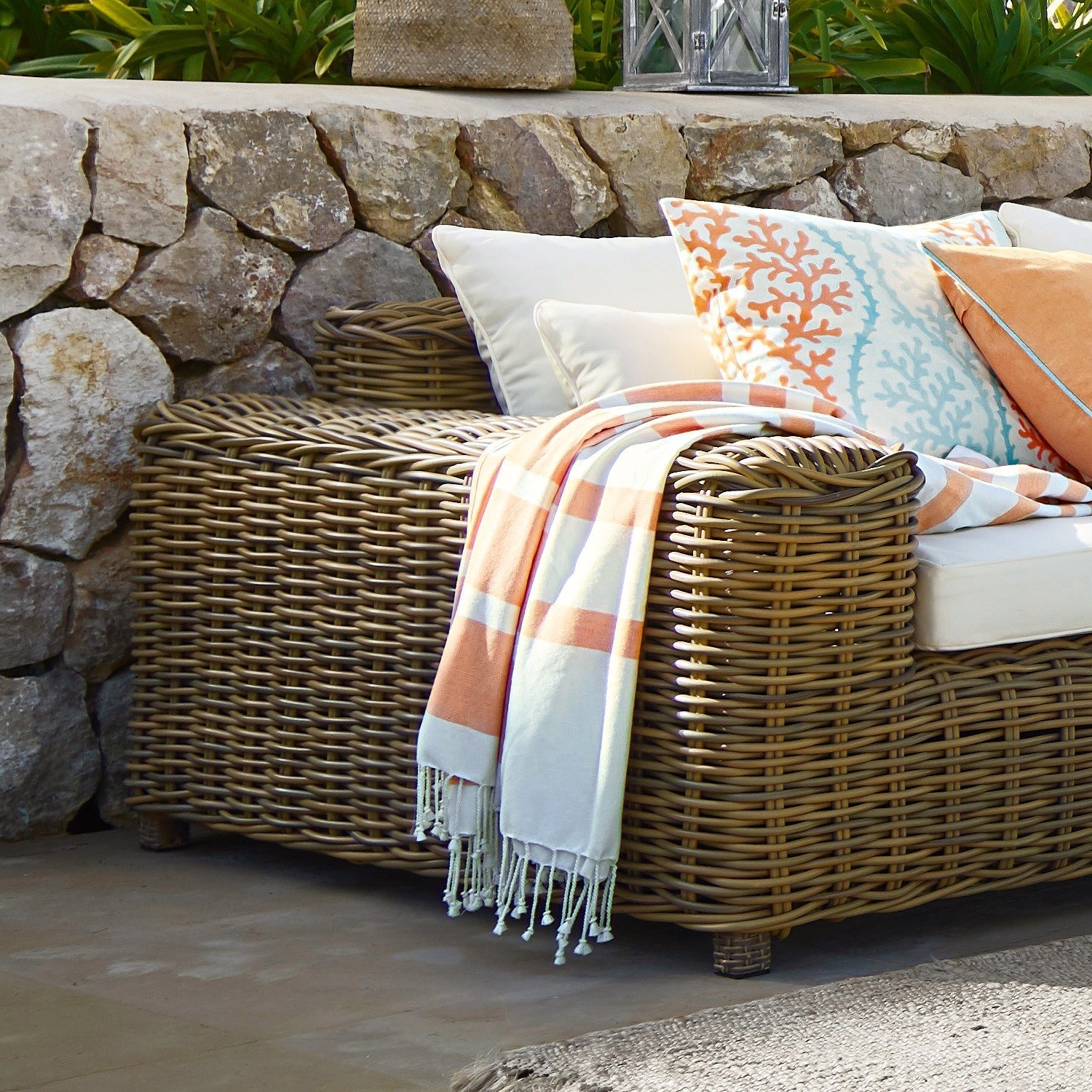 Braunes Rattan-Gartensofa mit cremefarbenen Kissen und orange-weißem Plaid auf Terrasse