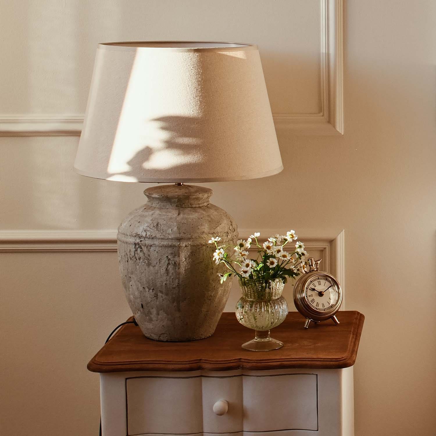 Tischlampe mit beigem Lampenschirm und vintage Keramikfuß auf Holztisch mit Blumen und Uhr