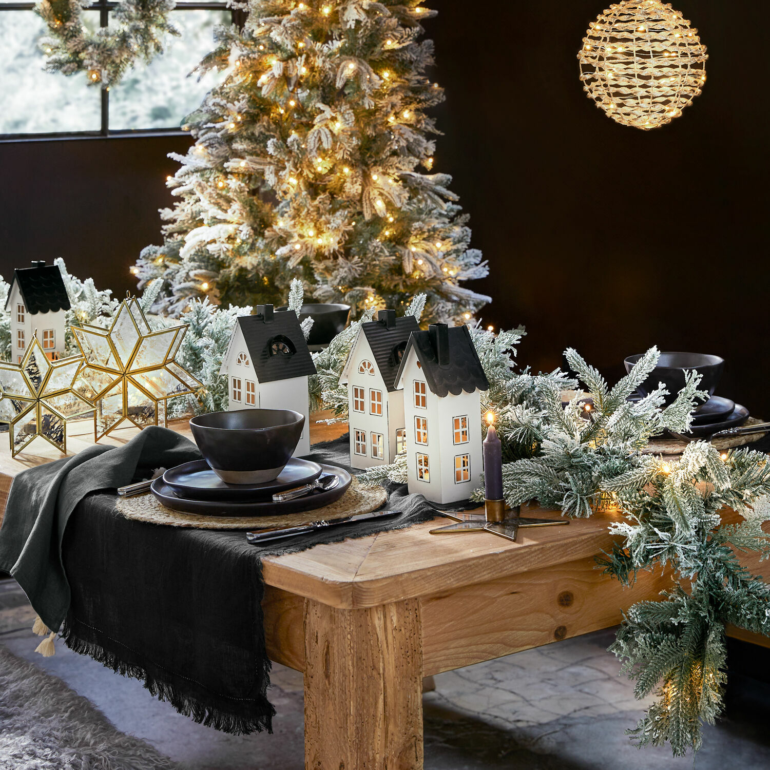 Weihnachtlich dekorierter Holztisch mit beleuchteten Häuschen, schneebedeckten Tannenzweigen und schwarzem Geschirr