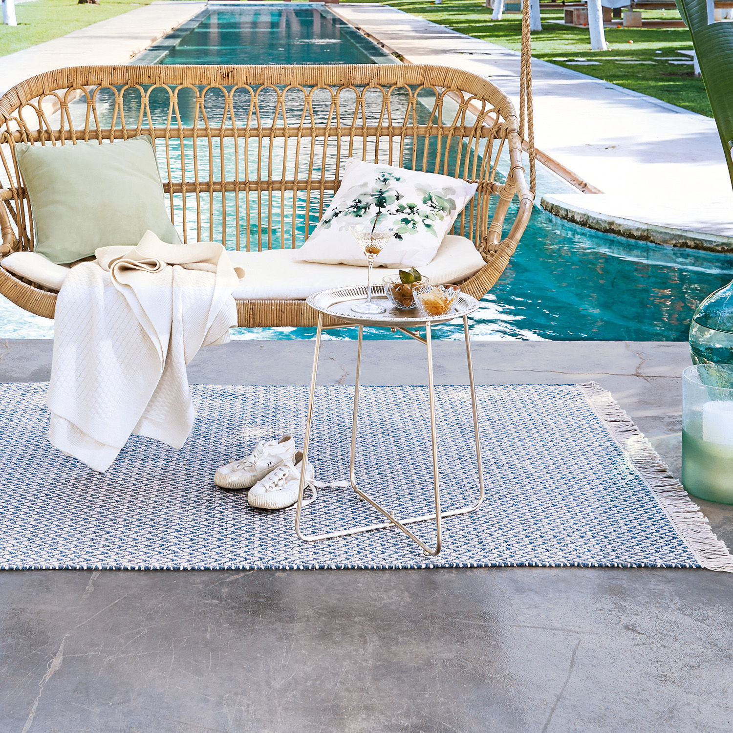 Hängesessel aus Natur-Rattan mit cremefarbenem Kissen und Beistelltisch am Pool auf blauem Teppich