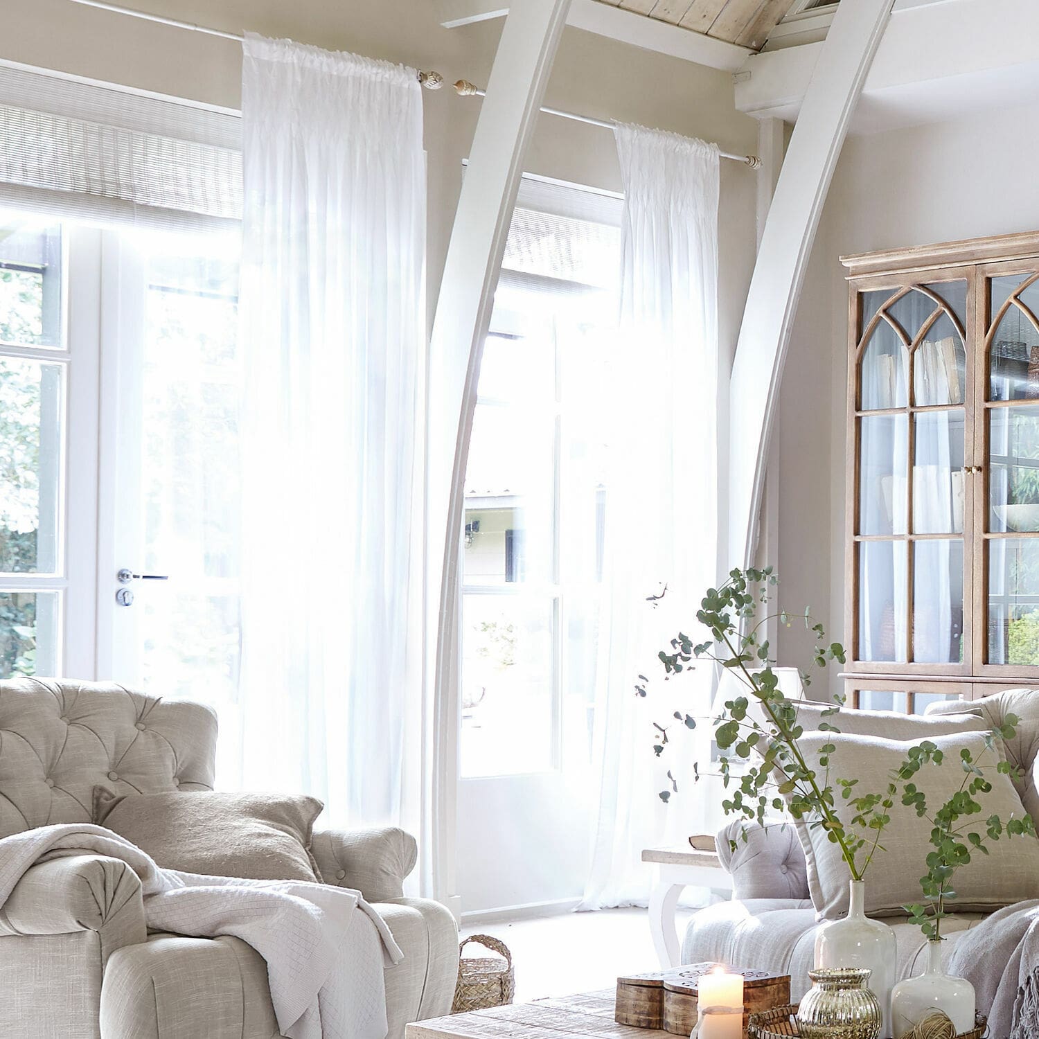 Gardine Otilie Helles Wohnzimmer mit beigem Polstersessel, weißen Vorhängen und vintage Holzvitrine