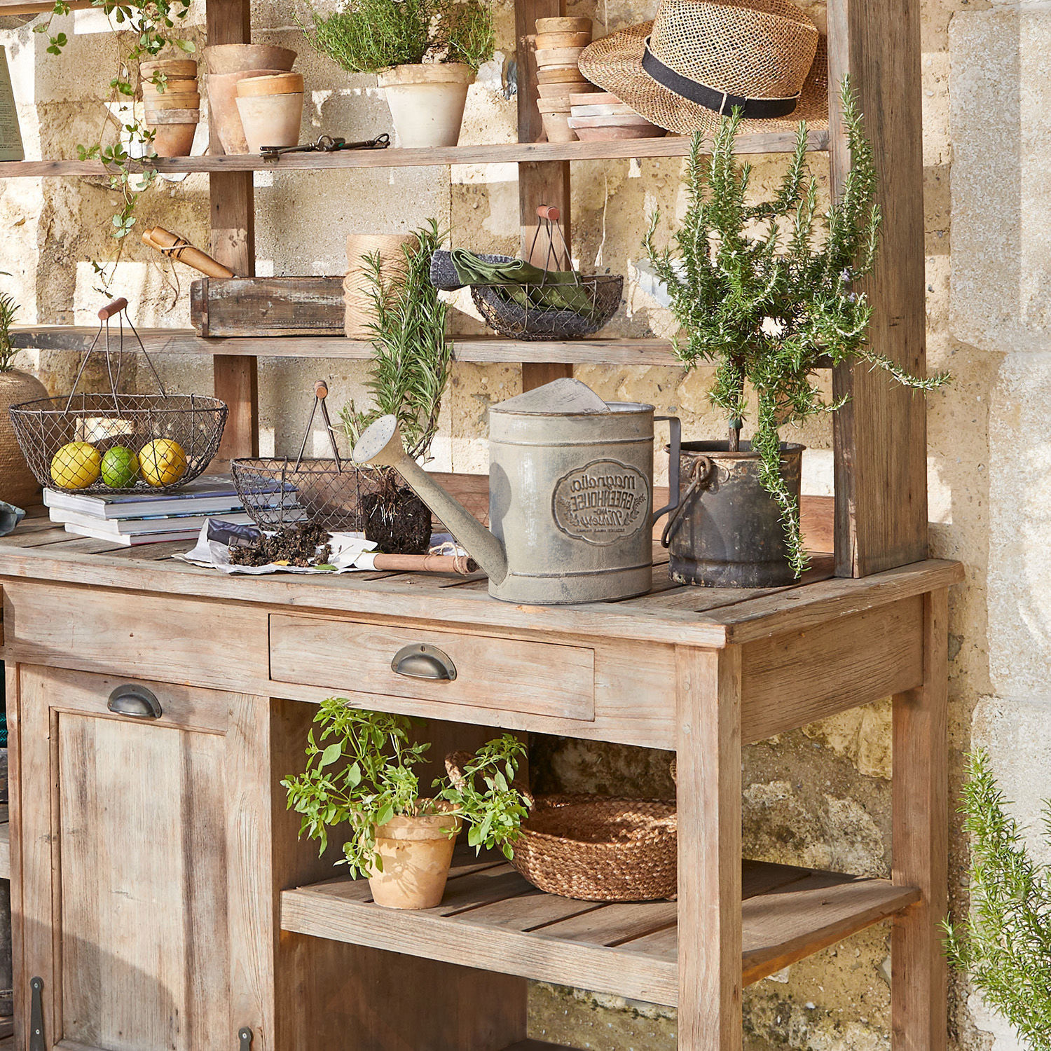 Holz-Gartentisch mit Metall-Gießkanne und Kräutertöpfen vor Steinmauer