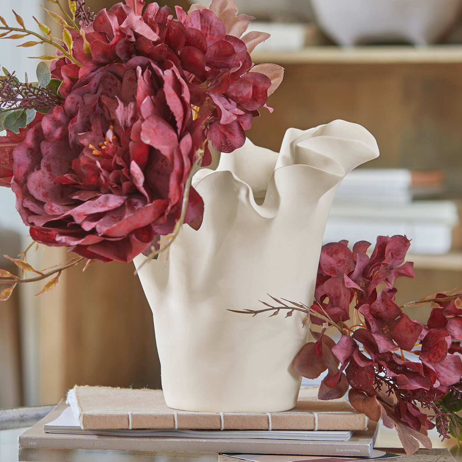 Vase Elvessa Weiße Keramikvase mit wellenförmigem Rand und roten künstlichen Blumen auf einem Tisch mit Büchern
