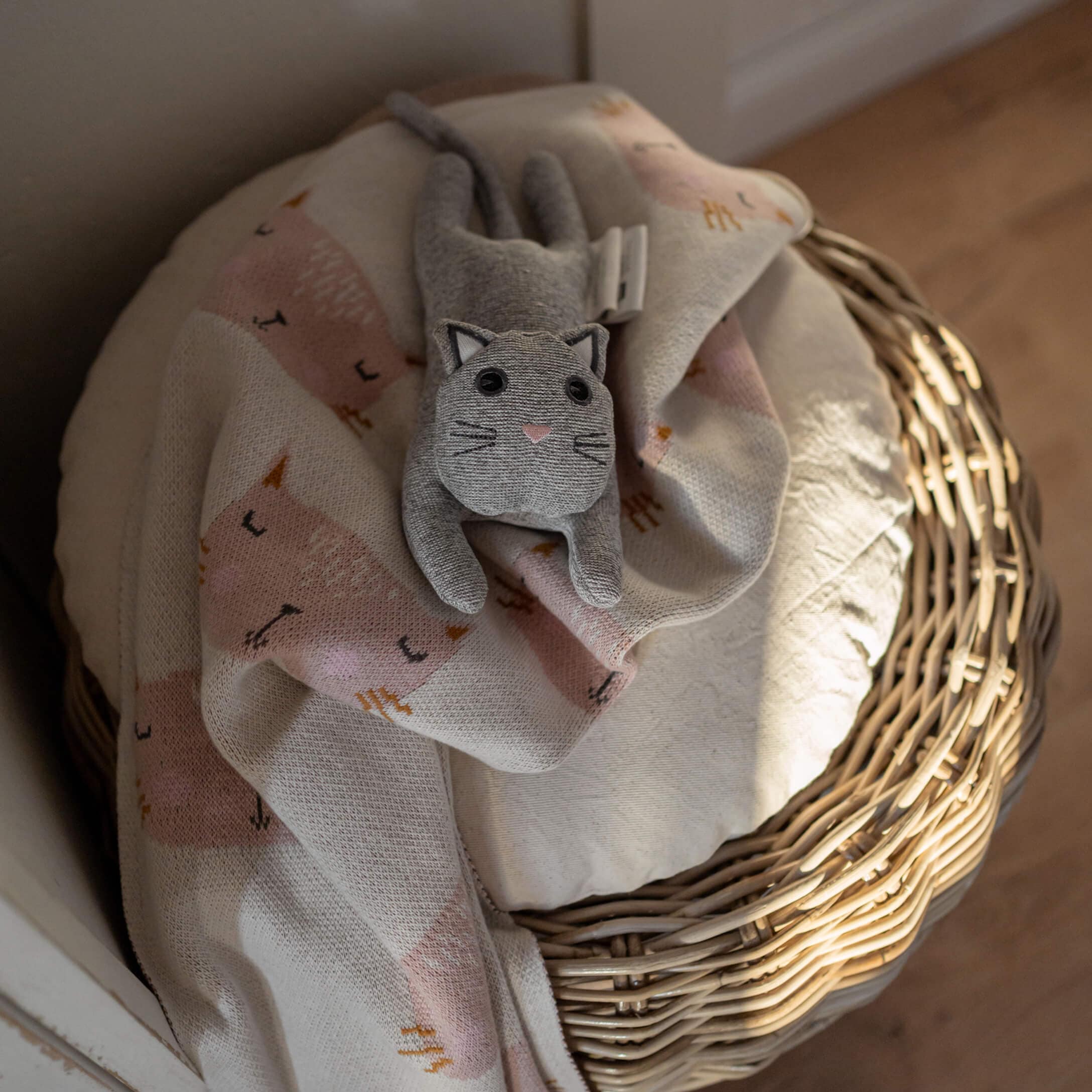 Graue Plüschkatze auf hellrosa Decke mit Katzenmotiv auf einem Weidenkorb