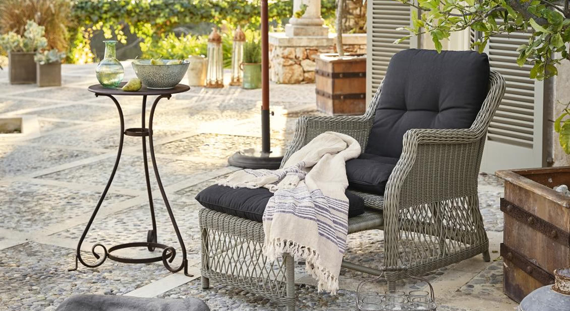 Grauer Rattansessel mit Fußhocker, schwarzen Kissen und beiger Decke auf steinerner Terrasse