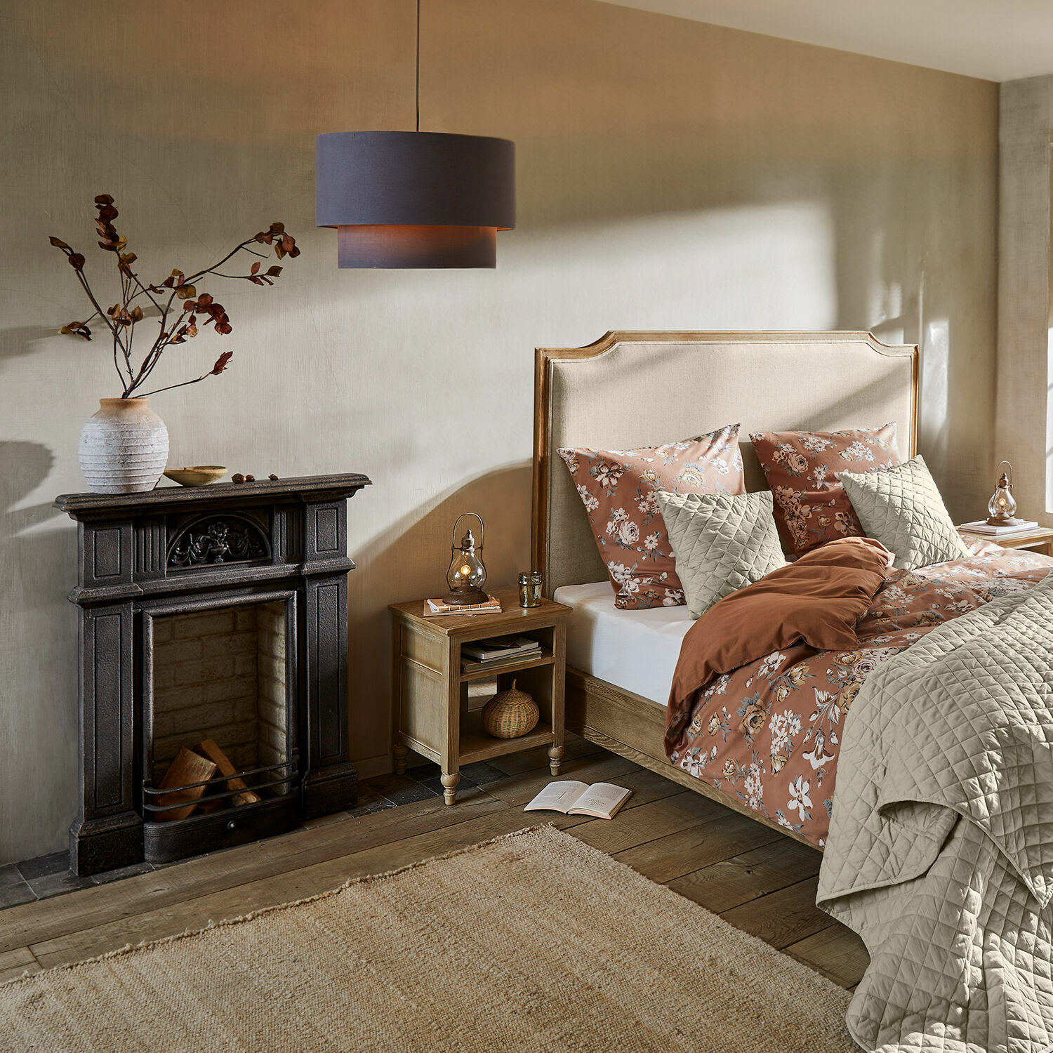 Gemütliches Schlafzimmer mit beige bezogenem Bett, brauner Blumenbettwäsche, Holzboden und dekorativem Kamin