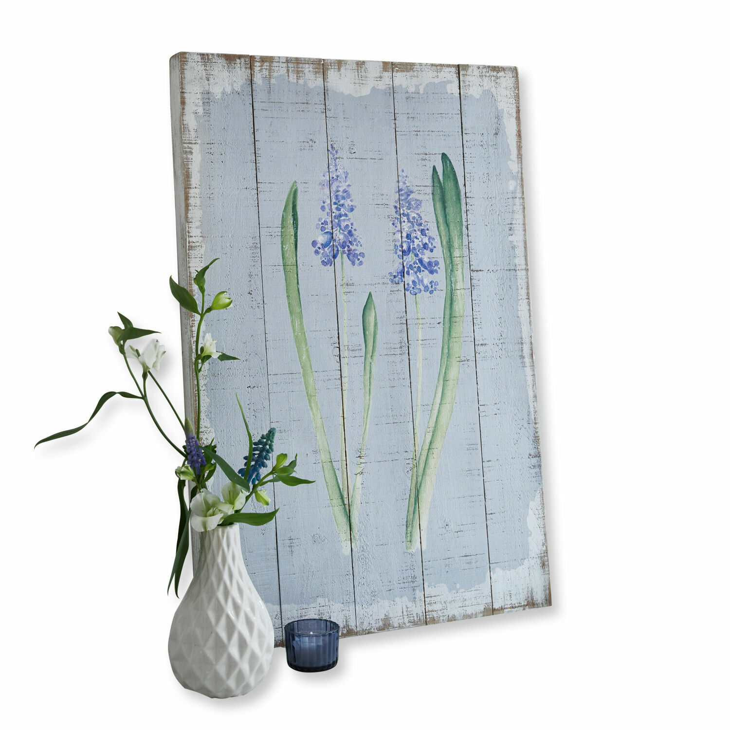 Holzbild mit blauem Blumenmotiv und weiße Keramikvase mit Blumen vor weißem Hintergrund