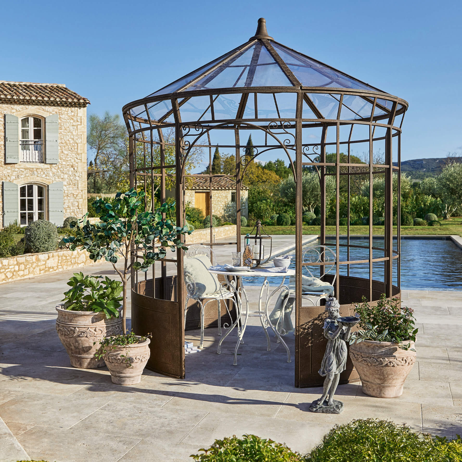 Garten mit rostfarbener Metalllaube, weißem Tisch und Stühlen, umgeben von Pflanzgefäßen