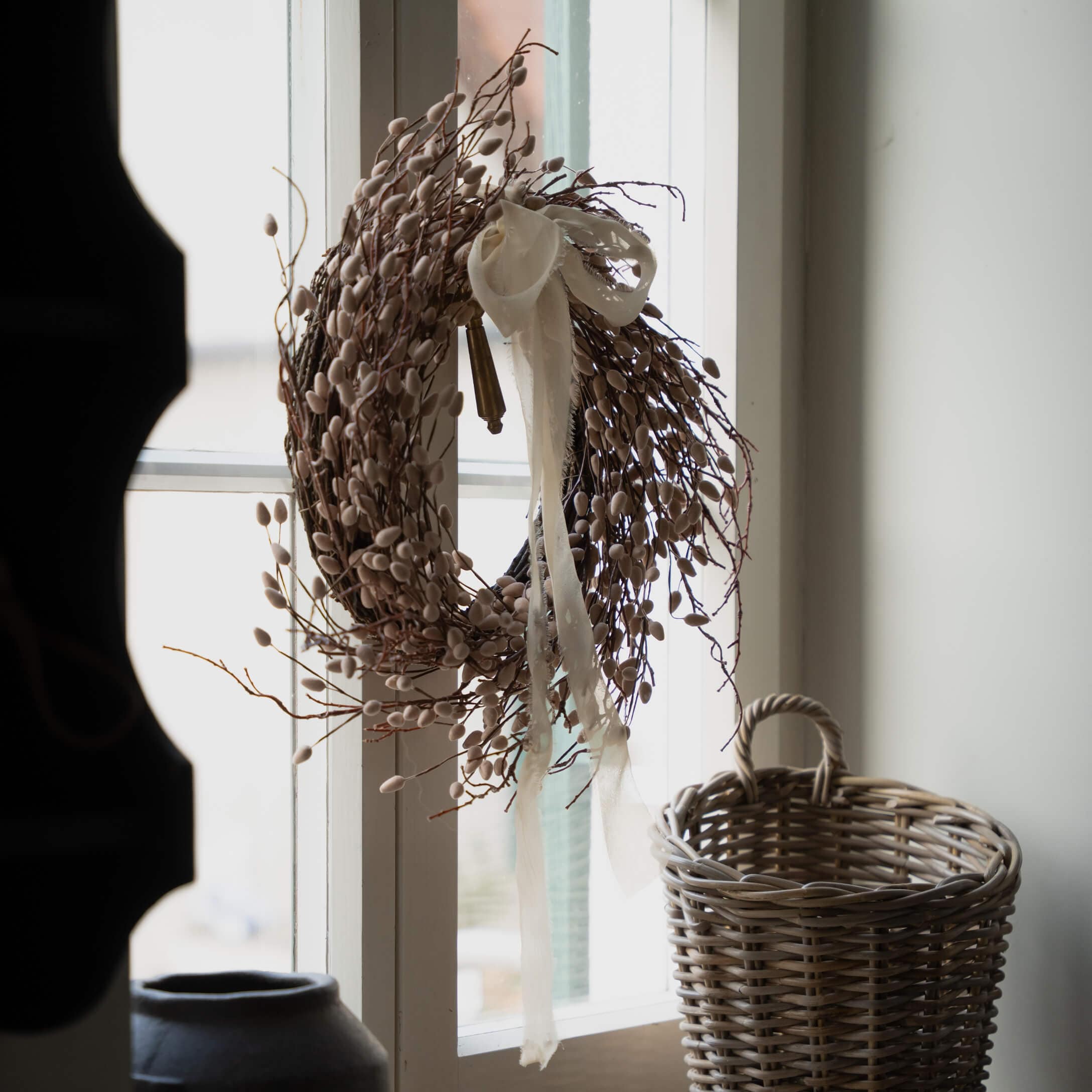 Dekorativer Kranz aus Zweigen und Knospen in Beige mit weißem Band am Fenster, daneben ein Weidenkorb
