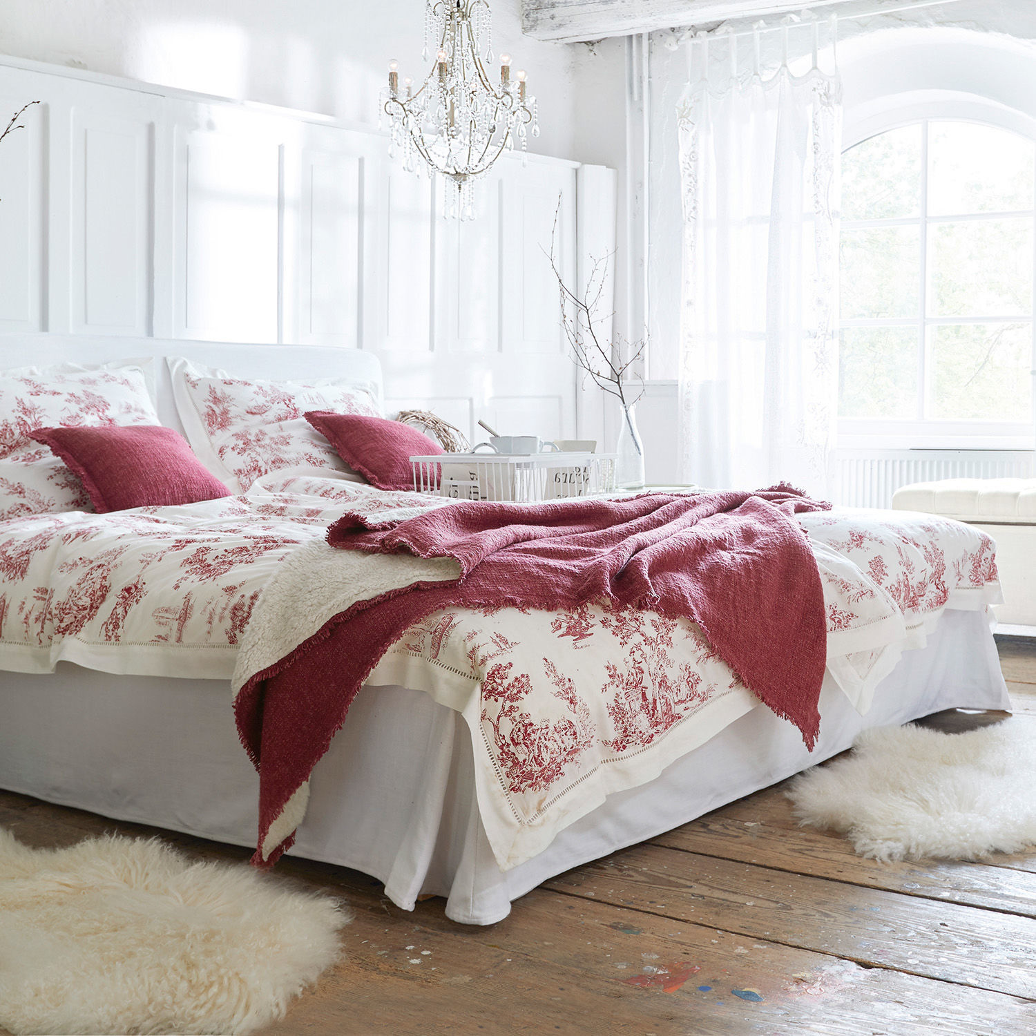 Schlafzimmer mit weißer Bettwäsche mit roten Mustern und bordeauxroter Decke auf Holzboden