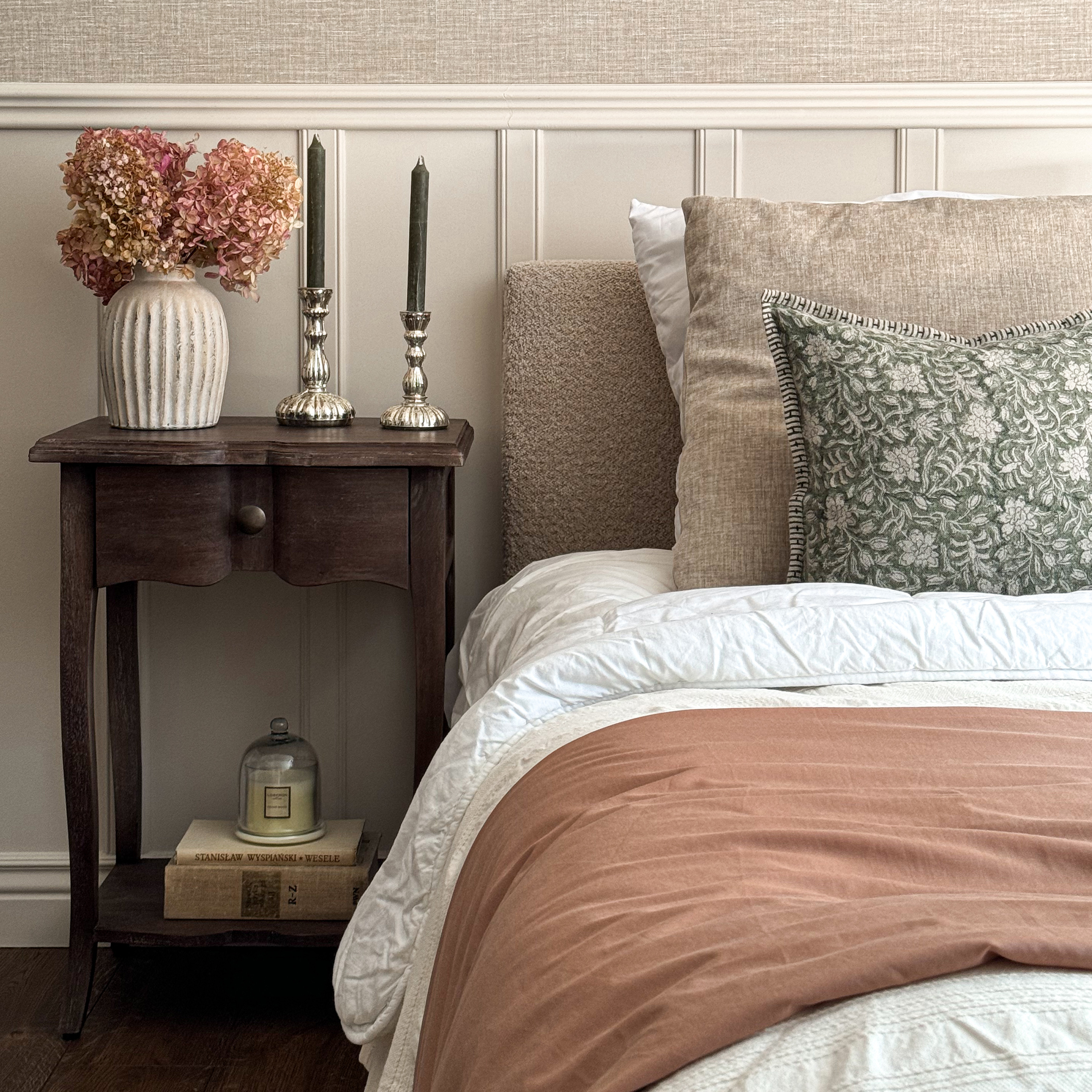 Gemütliches Schlafzimmer mit beiger Bettwäsche, grünem Kissen und Holznachttisch mit Dekorationen