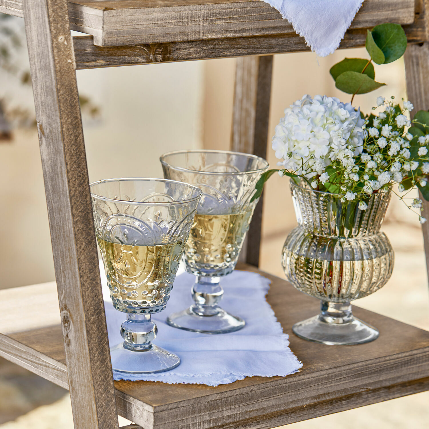 Zwei verzierte Gläser mit Weißwein und eine Vase mit weißen Blumen auf einem Holzregal