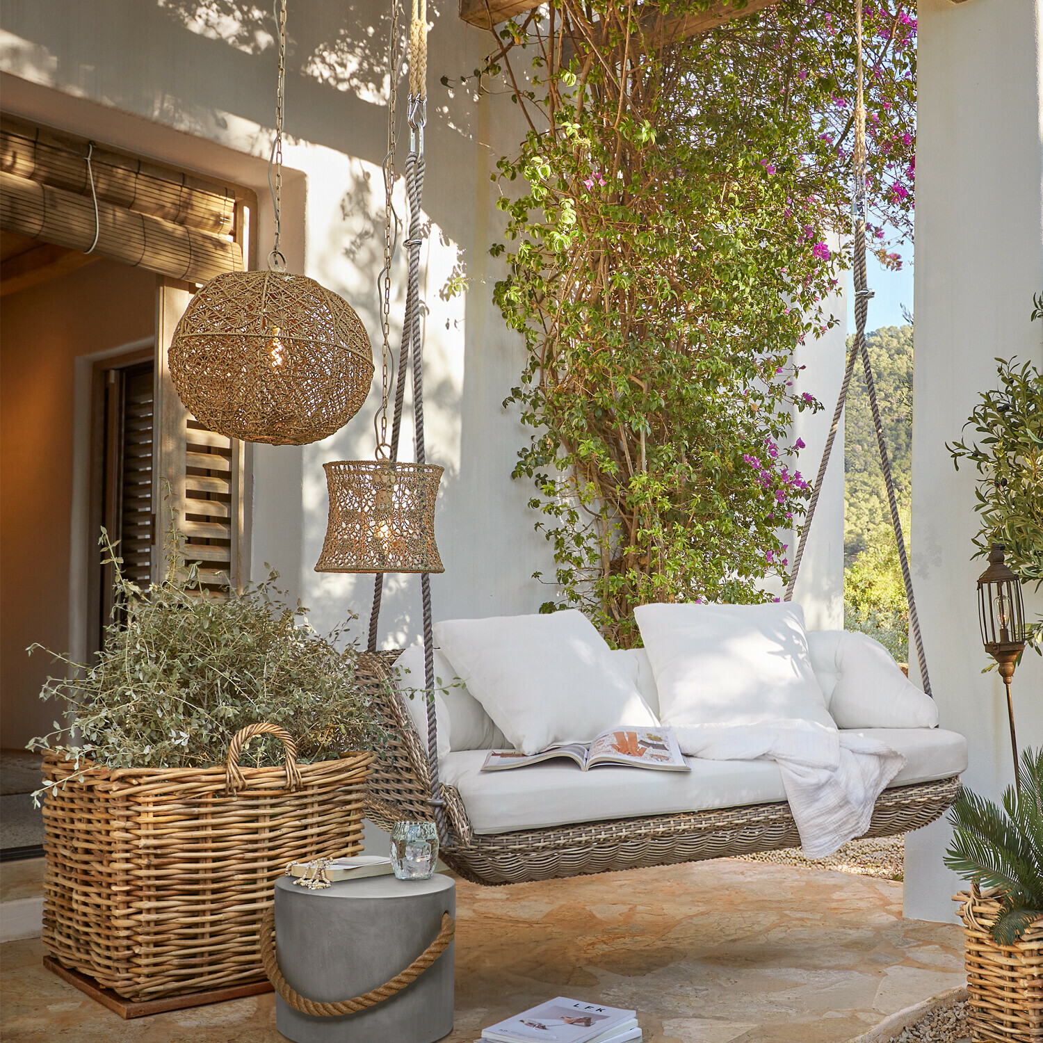 Rattanschaukel mit weißen Kissen auf Terrasse, Korb aus Weide und Lampen aus Naturmaterialien