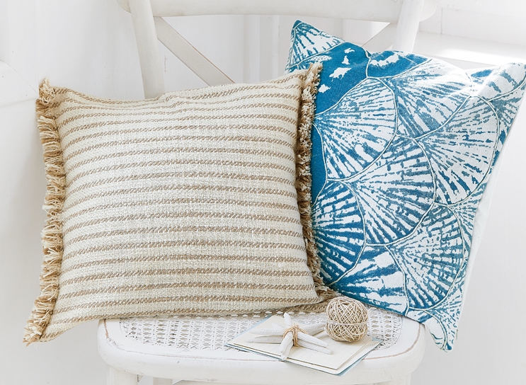 Zwei dekorative Kissen auf weißem Stuhl, eines beige gestreift mit Fransen, das andere blau mit Muschelmotiv