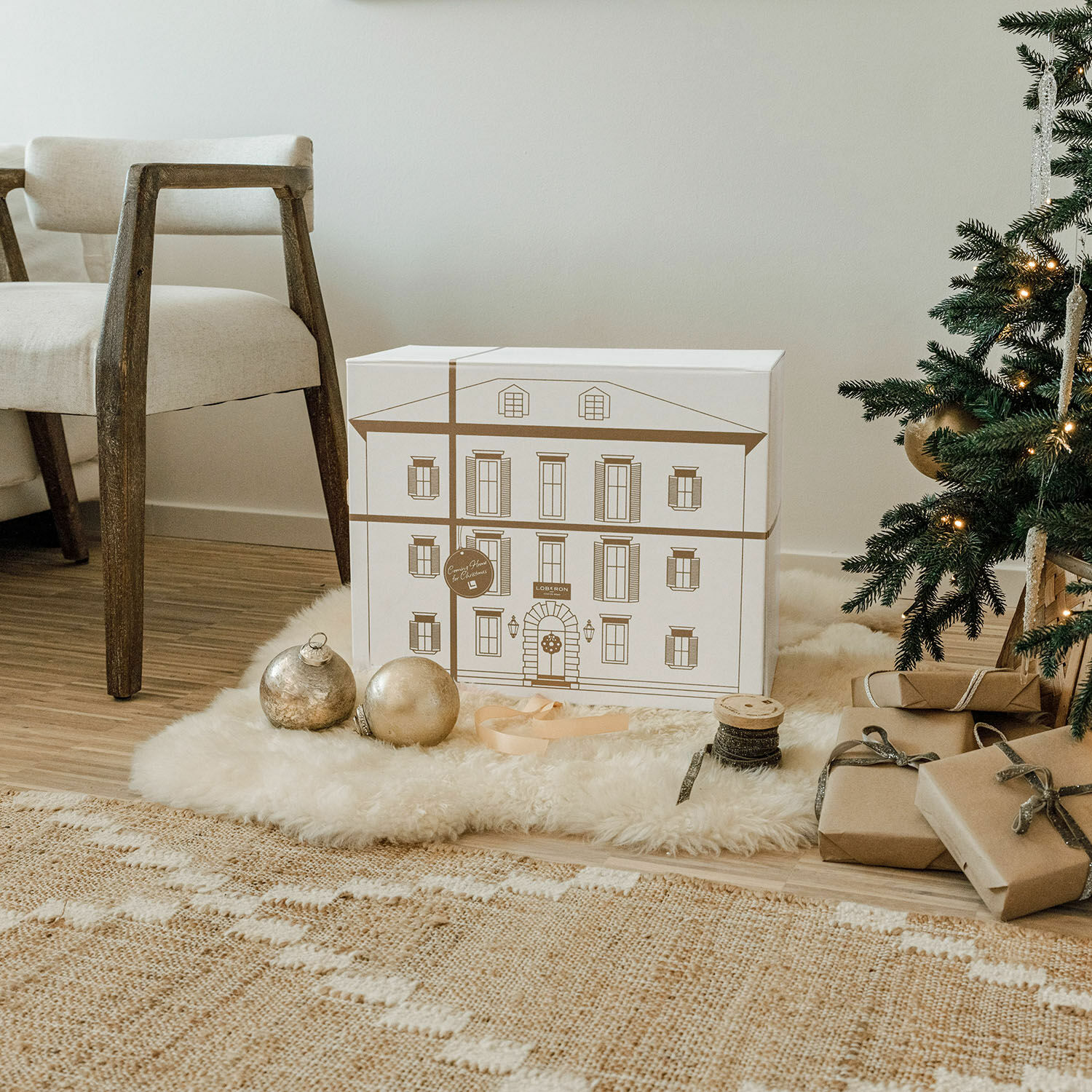 Dekorative Geschenkbox mit Hausmotiv auf weißem Fell neben Weihnachtsbaum und Naturgeschenken