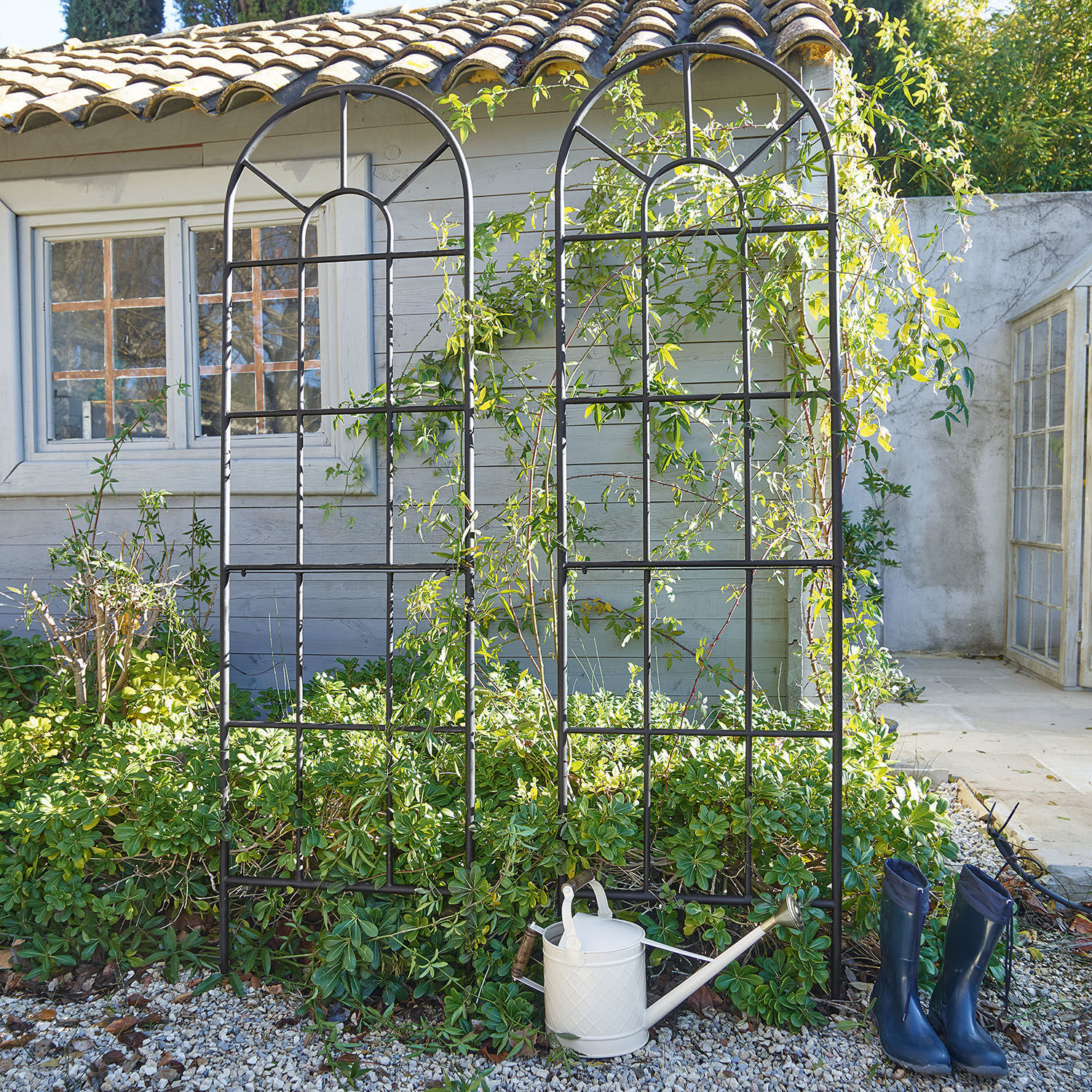 Rankgitter 2er Set Ealdred Schwarzes Metall-Gartenrankgitter mit Bogenmuster vor grünem Bewuchs und Haus, Gießkanne und Gummistiefel