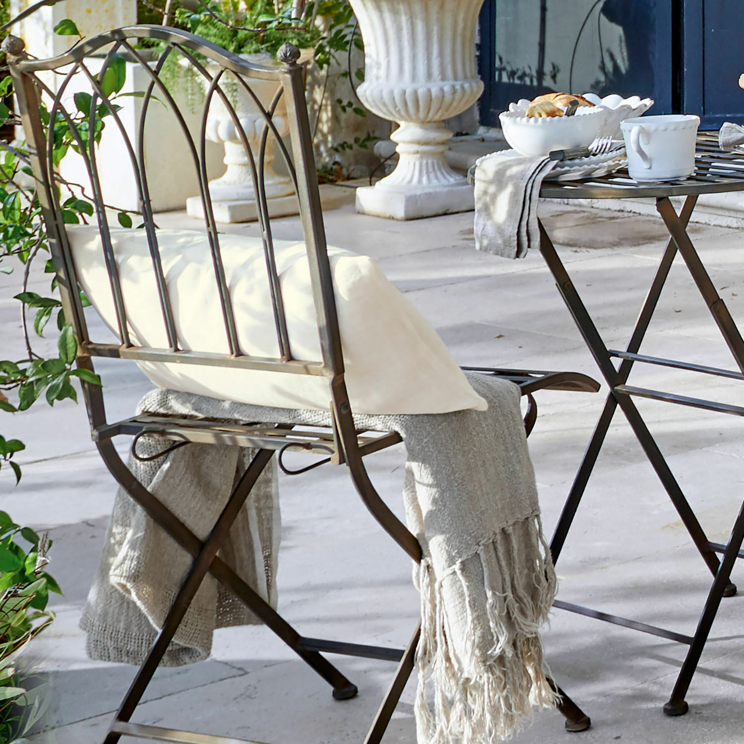 Metall-Gartenstuhl mit weißem Kissen und grauem Plaid auf heller Stein-Terrasse
