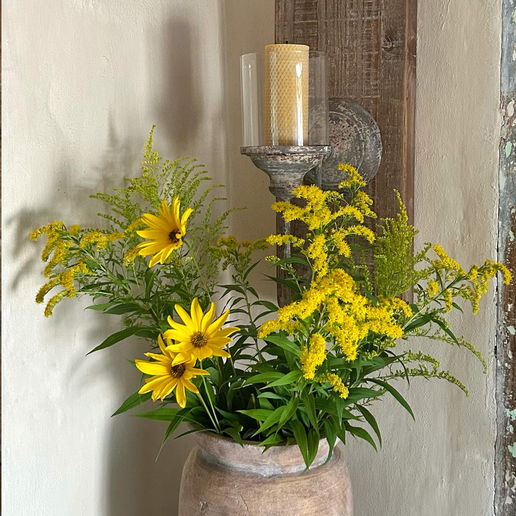 Gelbe Blumen in einem Tonkrug vor einer Wand mit rustikalem Kerzenhalter