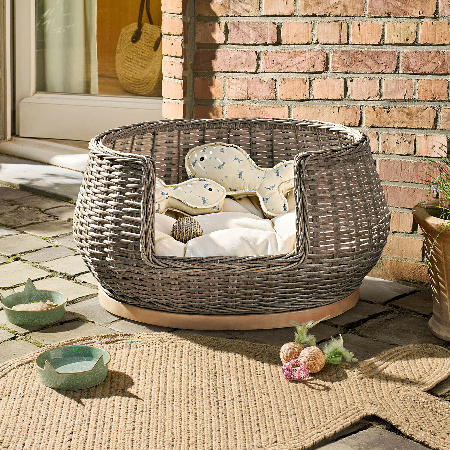 In- & Outdoor-Haustierbett Brinelza Rundes Hundebett aus grauem Geflecht mit cremefarbenem Kissen und Spielzeug auf Steinboden
