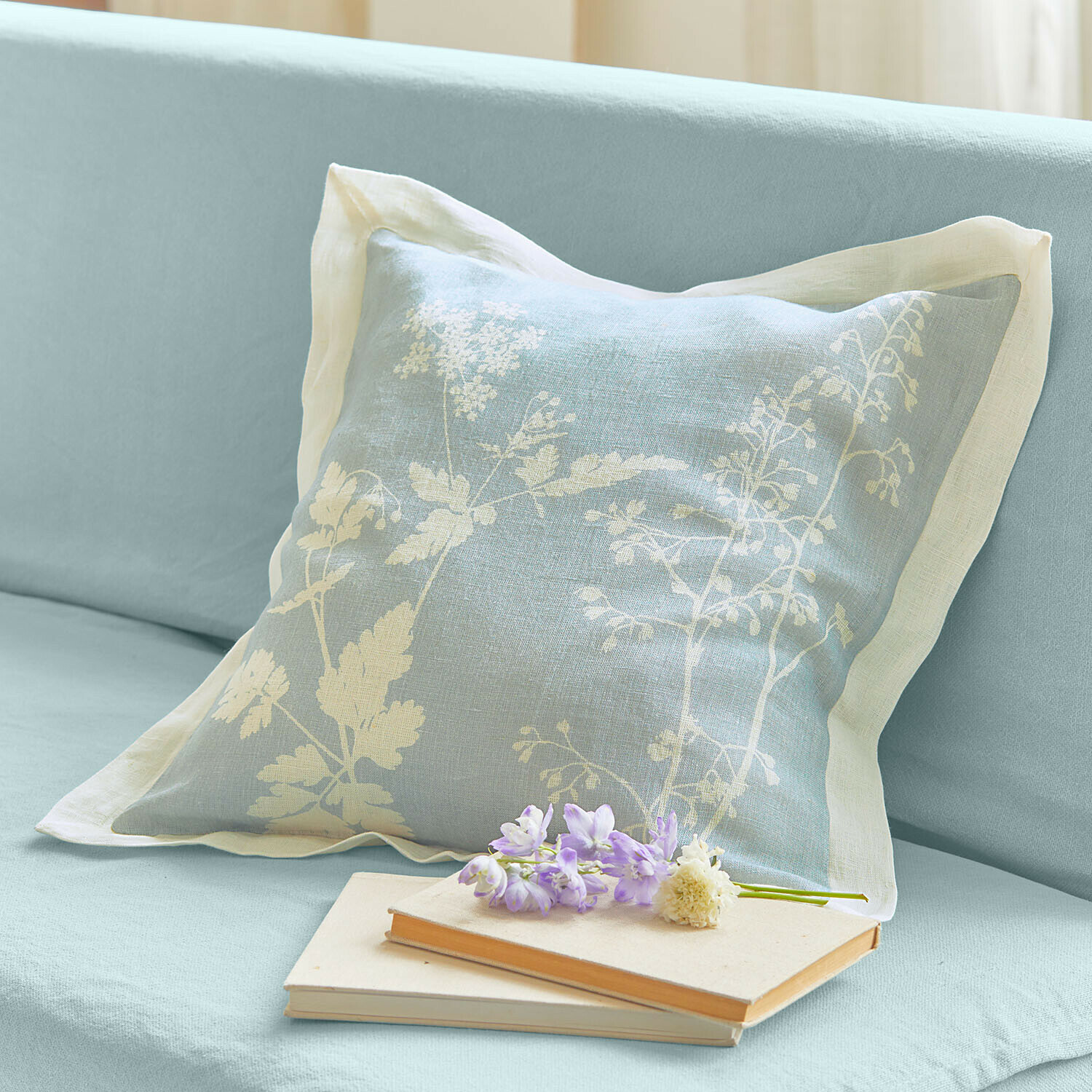 Kissenhülle Hazelwick Hellblaues Leinenkissen mit weißem Blumenmuster auf Sofa, daneben Bücher und Blumen