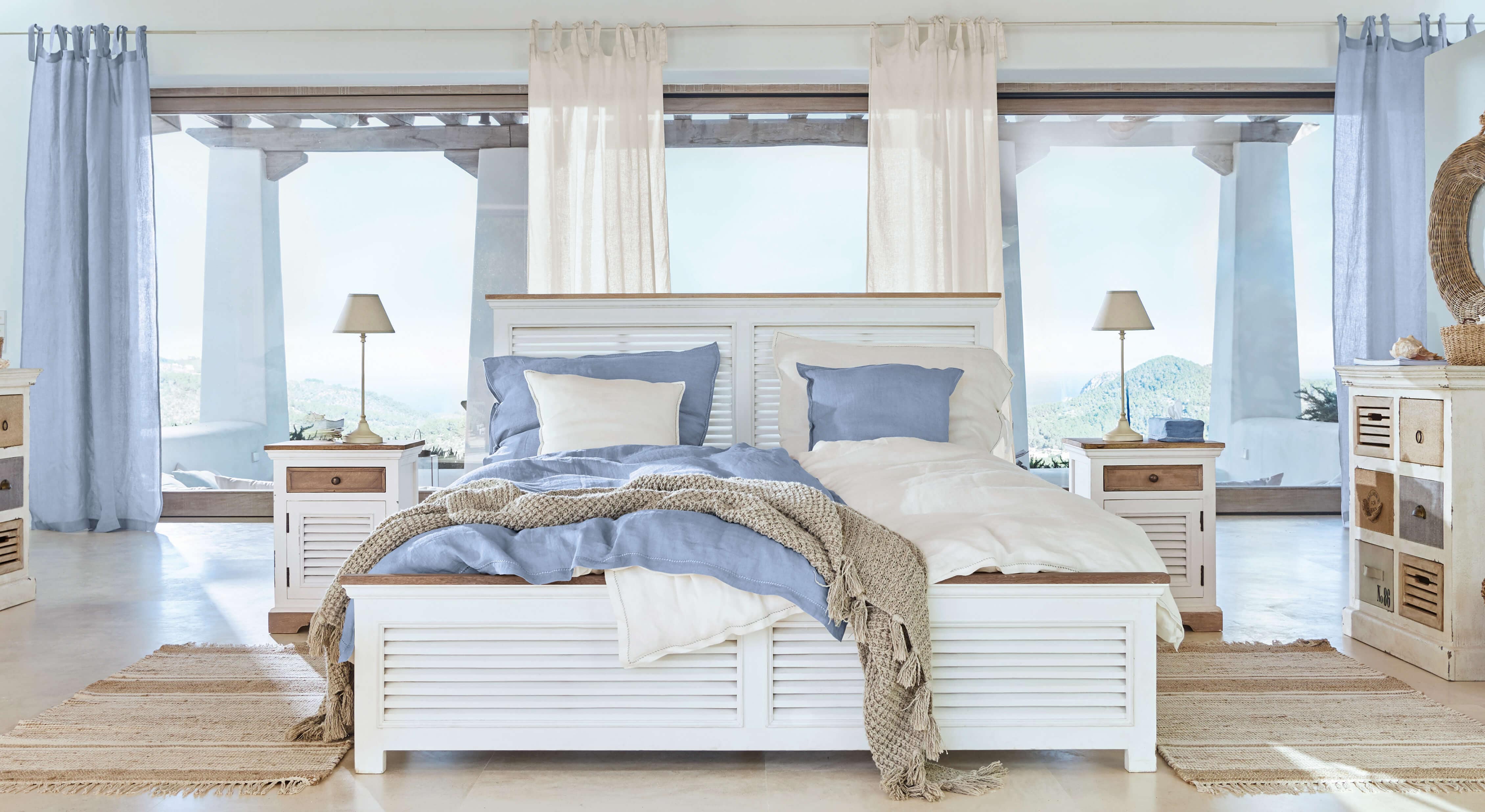 Weißes Holzbett mit blau-beiger Bettwäsche und Naturteppich in hellem Schlafzimmer mit Aussicht