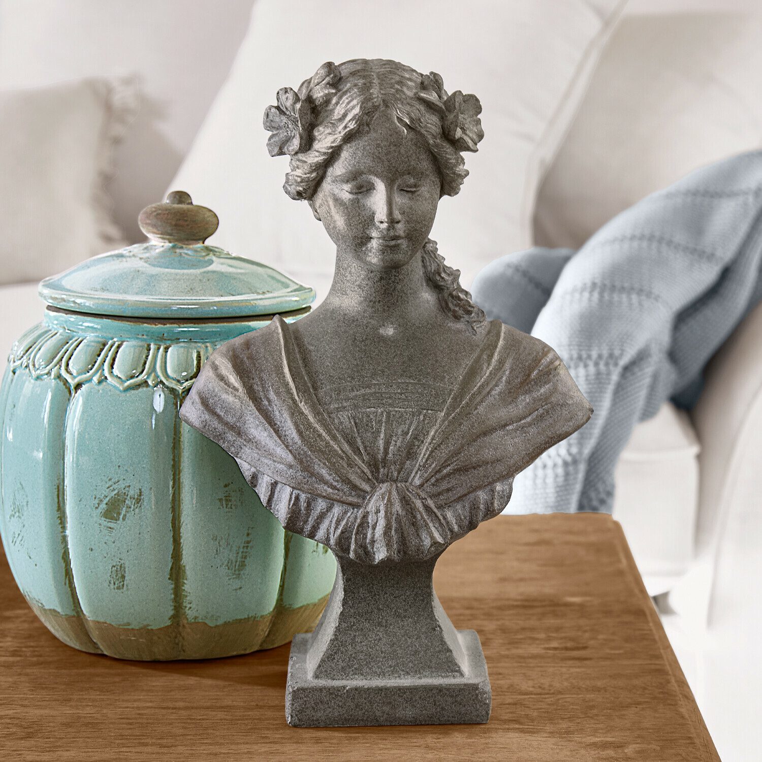 Graue Steinstatue einer Frau mit Blumen im Haar auf Holztisch neben türkisfarbenem Keramikgefäß