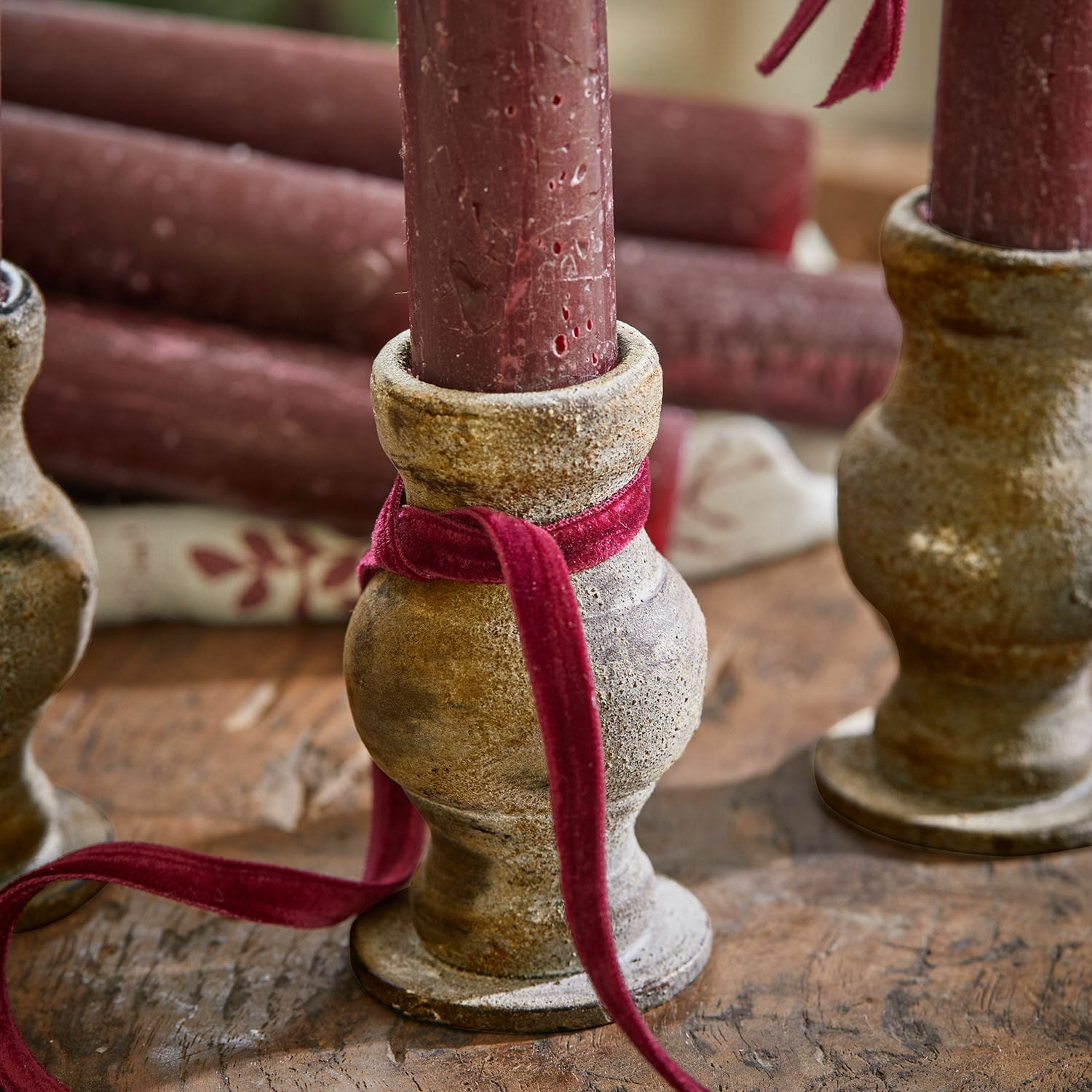 Rustikaler Keramik-Kerzenhalter in Beige mit bordeauxroter Kerze und Samtband auf Holztisch