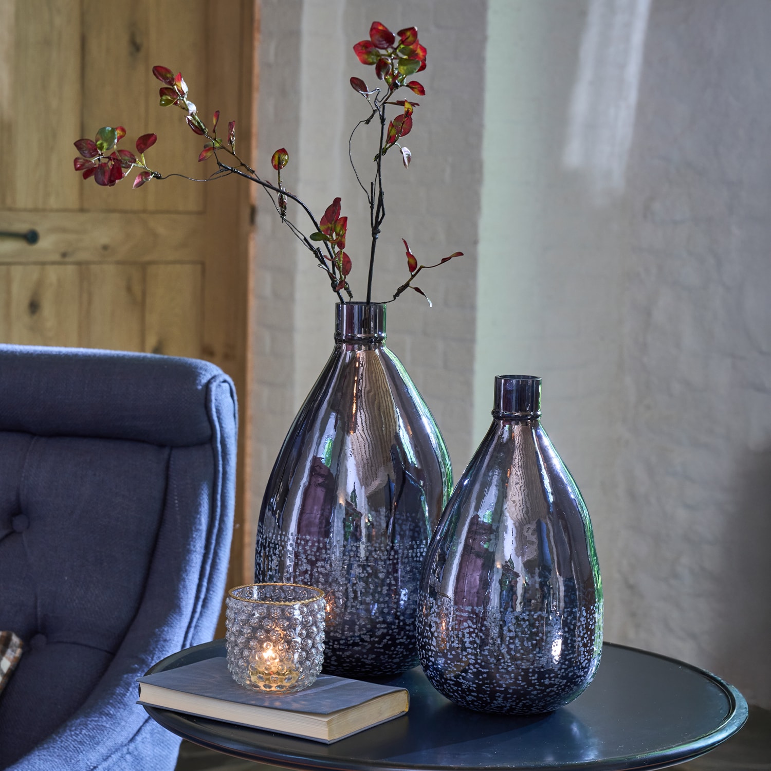 Zwei dekorative dunkelgraue Glasvasen mit roten Zweigen auf einem Tisch neben Kerzenhalter und Buch