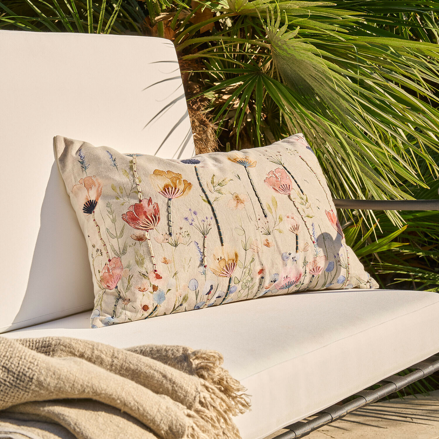 Beiges Dekokissen mit Blumenmuster auf hellem Gartenstuhl mit Decke