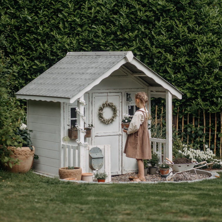 Weißes Holz-Gartenhaus mit dekorativem Kranz und Pflanzen, Kind hält Blumentopf