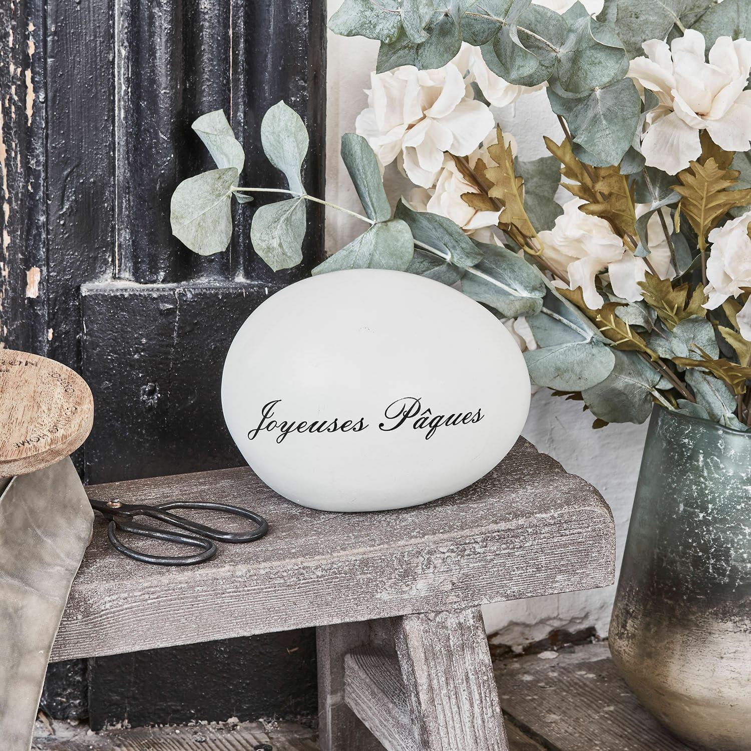 Deko-Ei JOYEUSES PAQUES Weißes Dekoei mit Schriftzug Joyeuses Pâques auf Holzbank mit Blumen und Schere