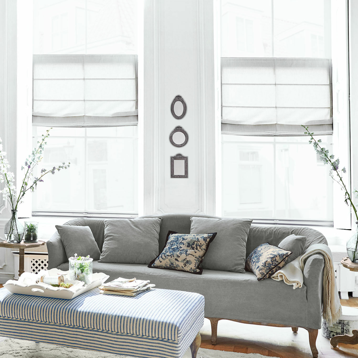 Graues Stoffsofa mit Kissen und blau-weiß gestreiftem Hocker in hellem Wohnzimmer mit Dekoration