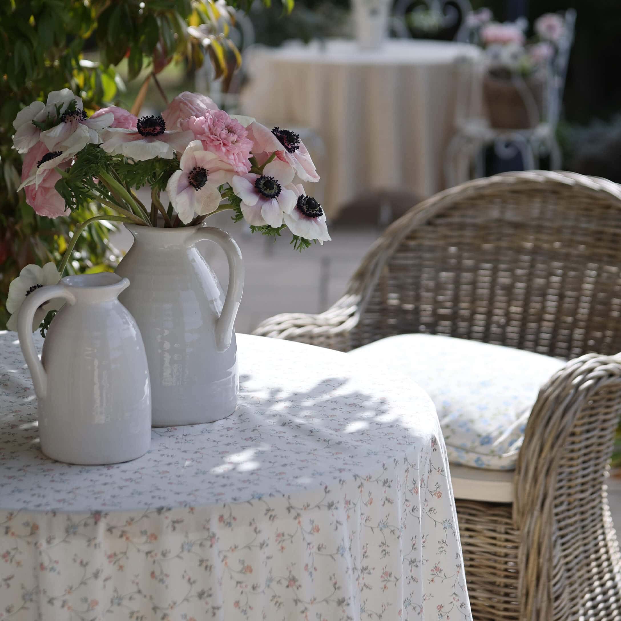 Weiße Keramikkrüge mit rosa und weißen Blumen auf einem Tisch mit floralem Tischtuch im Garten