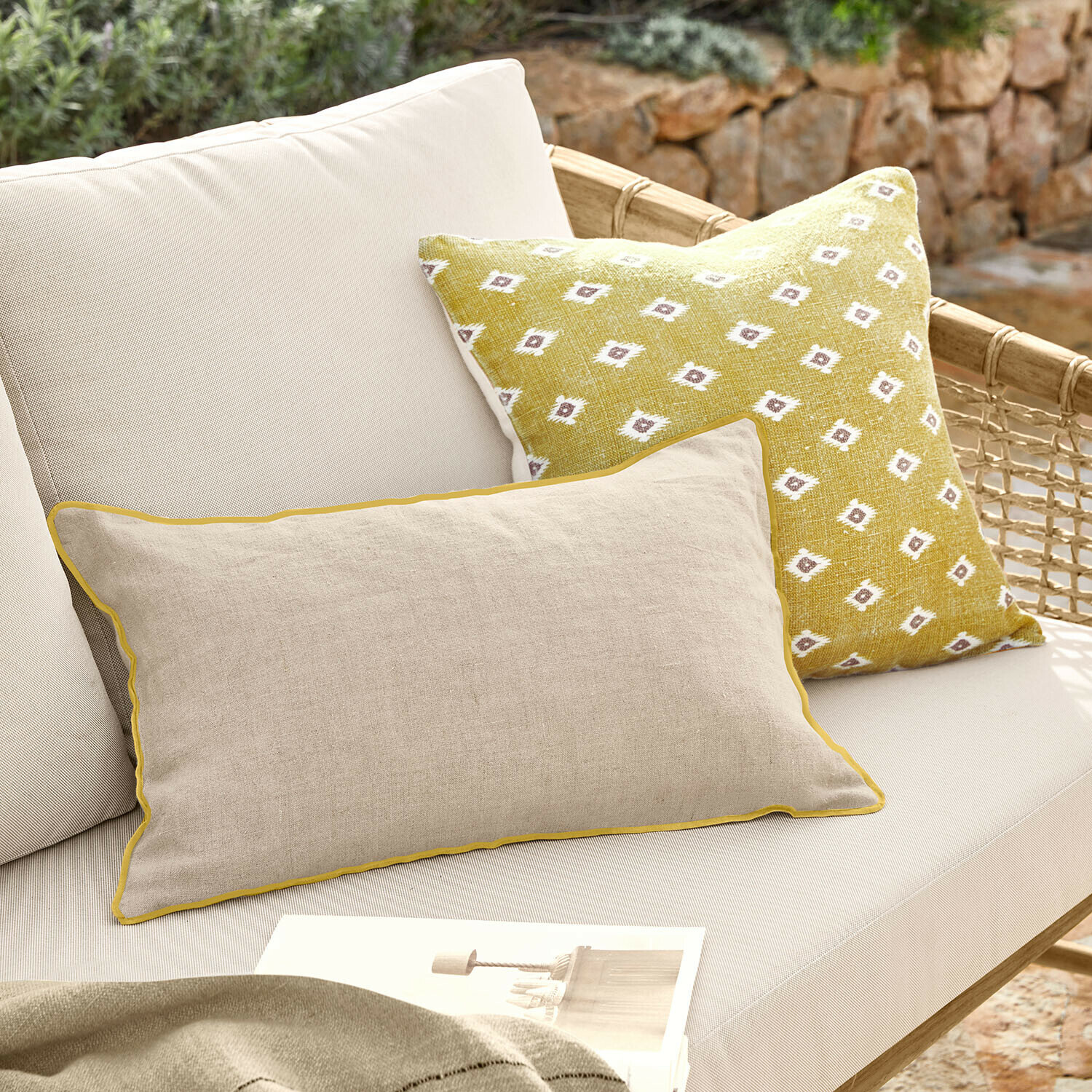 Beiges Leinenkissen mit gelber Paspel und gelbes Kissen mit weißem Muster auf hellem Gartensofa