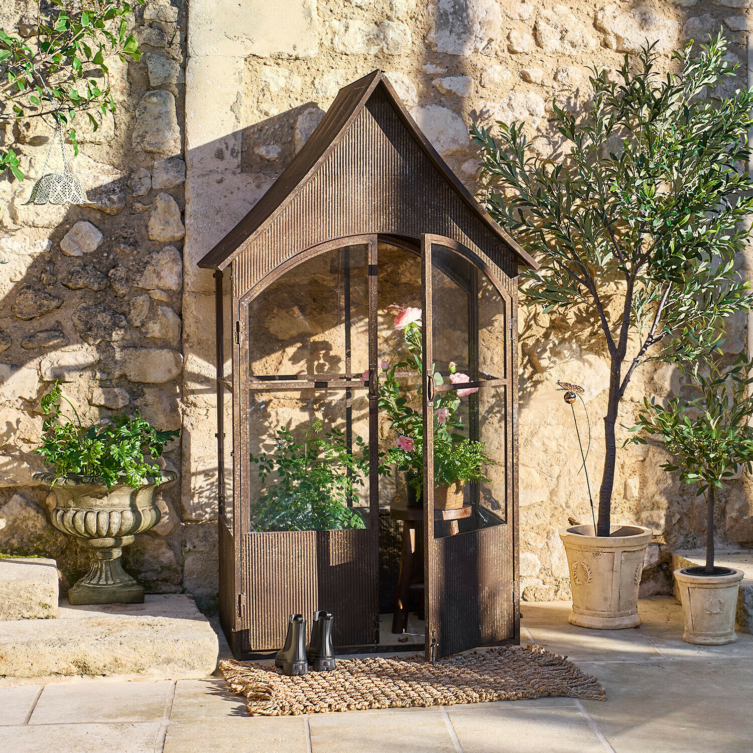 Vintage Metall-Gewächshaus in Braun mit Pflanzen und Töpfen vor sonniger Steinmauer im Garten