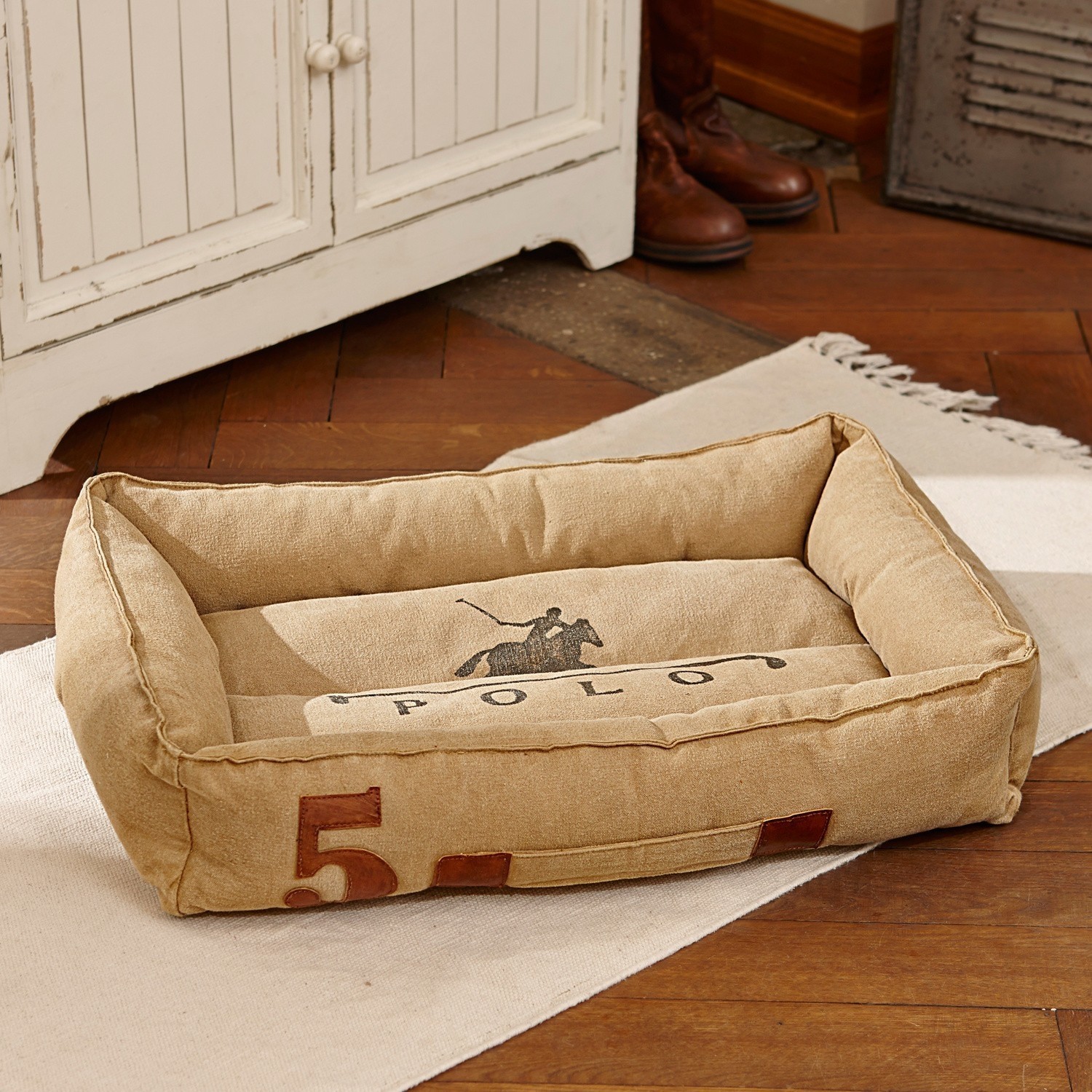 Beiges Hundebett aus robustem Material mit Polo-Motiv und Zahl 5, auf Holzboden platziert.