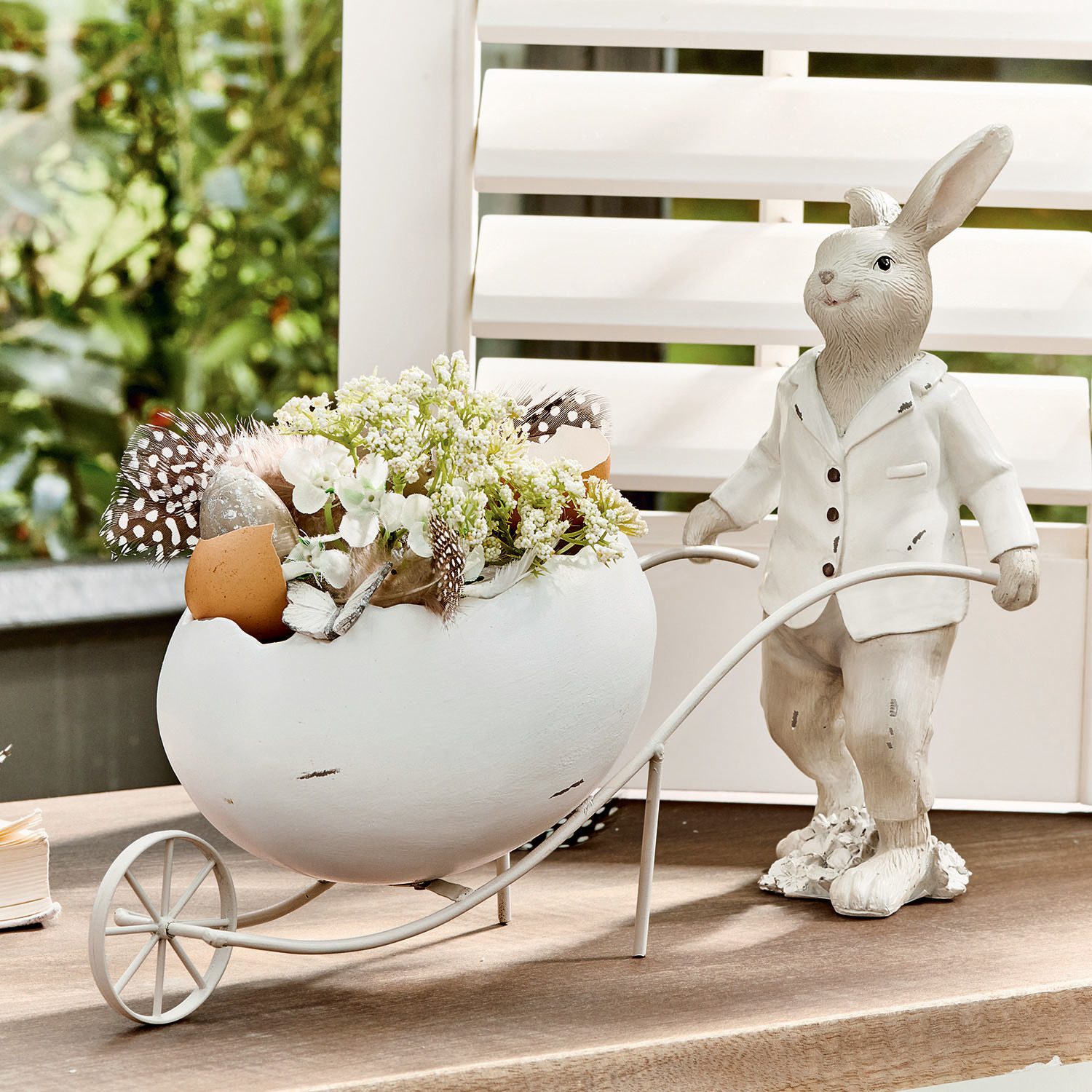 Hase Jaris Dekorativer weißer Hase mit Keramikei und Blumen in Metallwagen auf Tisch.