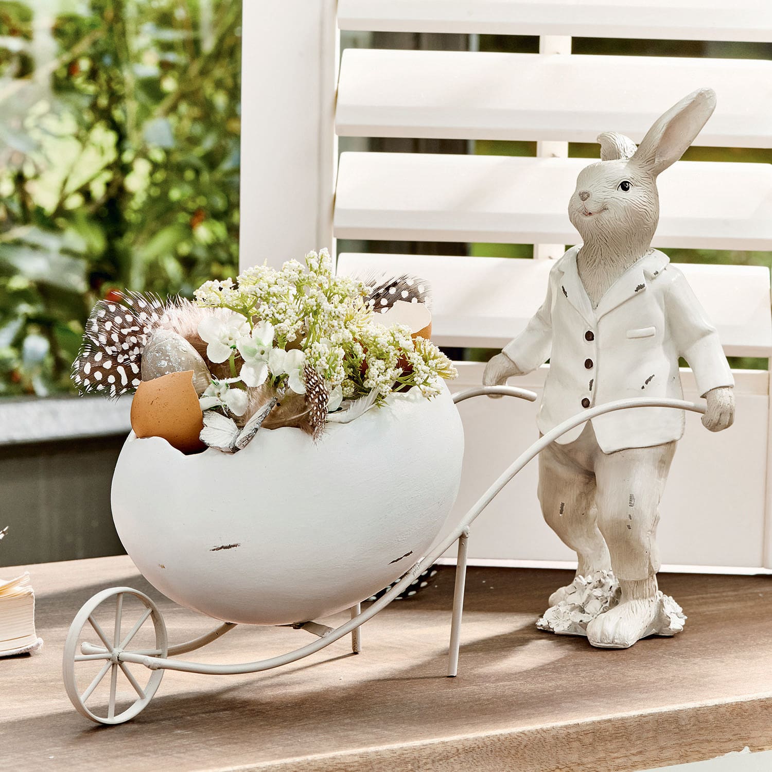 Hase Jaris Dekorativer weißer Hase mit Keramikei und Blumen in Metallwagen auf Tisch.