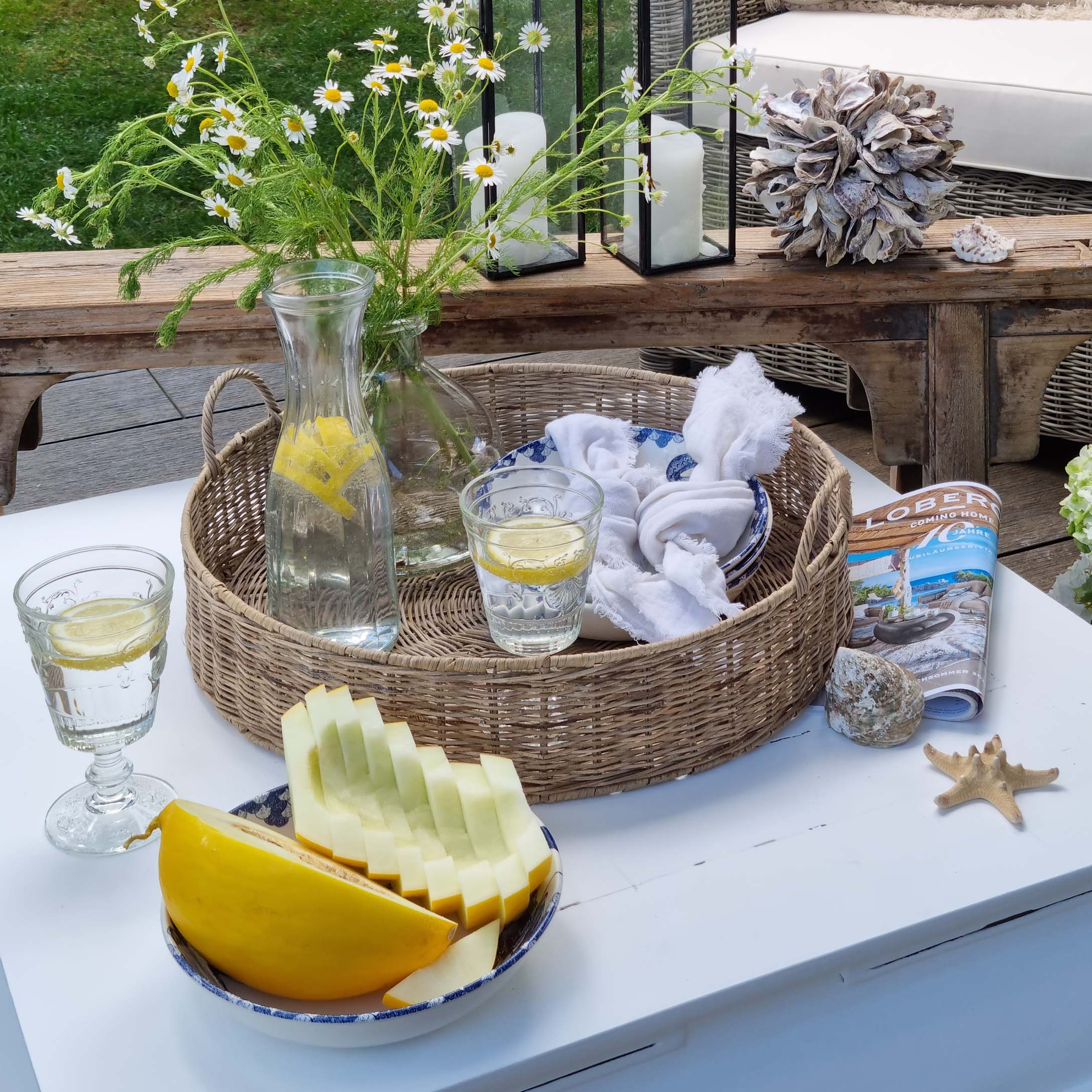 Runder Korb aus Rattan mit Zitronenwasser und Glas auf weißem Gartentisch dekoriert