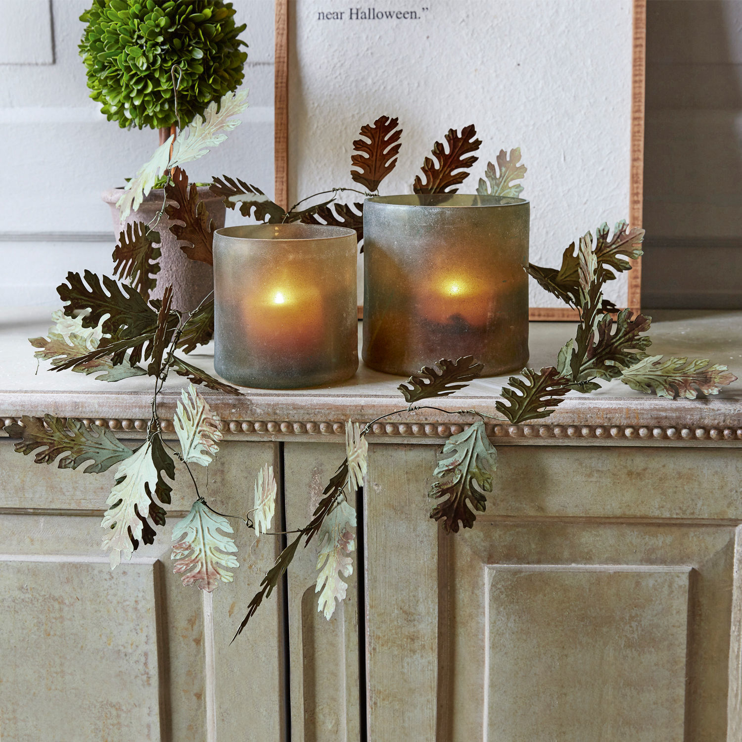 Zwei Glas-Teelichthalter mit grün-braunen Metallblättern auf beigem Schrank, Wohnaccessoire