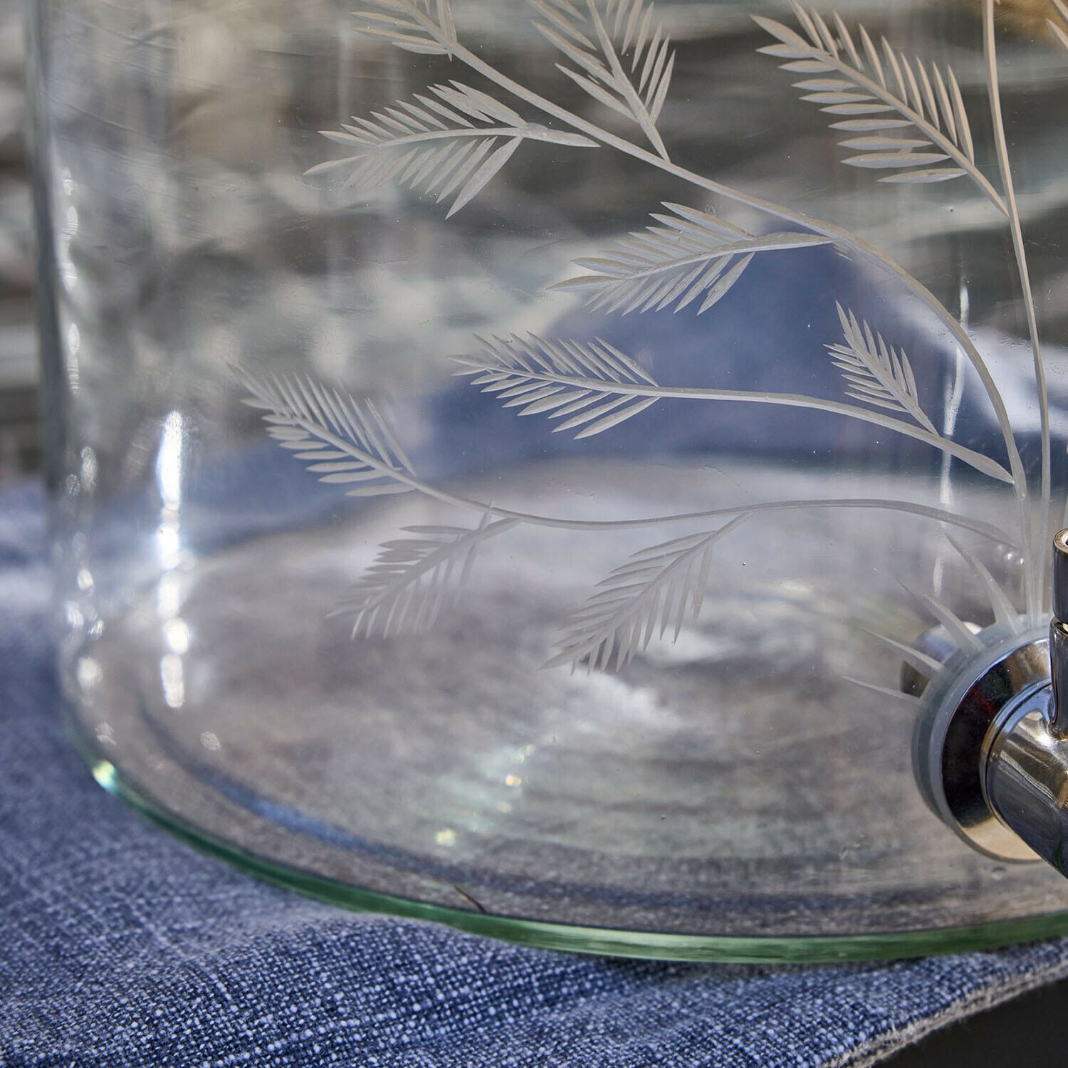 Glasbehälter mit graviertem Blattmuster und Metallhahn auf blauem Stoff