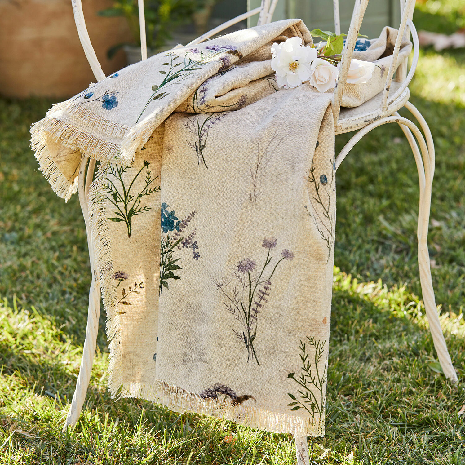 Plaid Cabinda Beige Leinentischdecke mit Blumenmuster und Fransen auf weißem Metallstuhl im Garten
