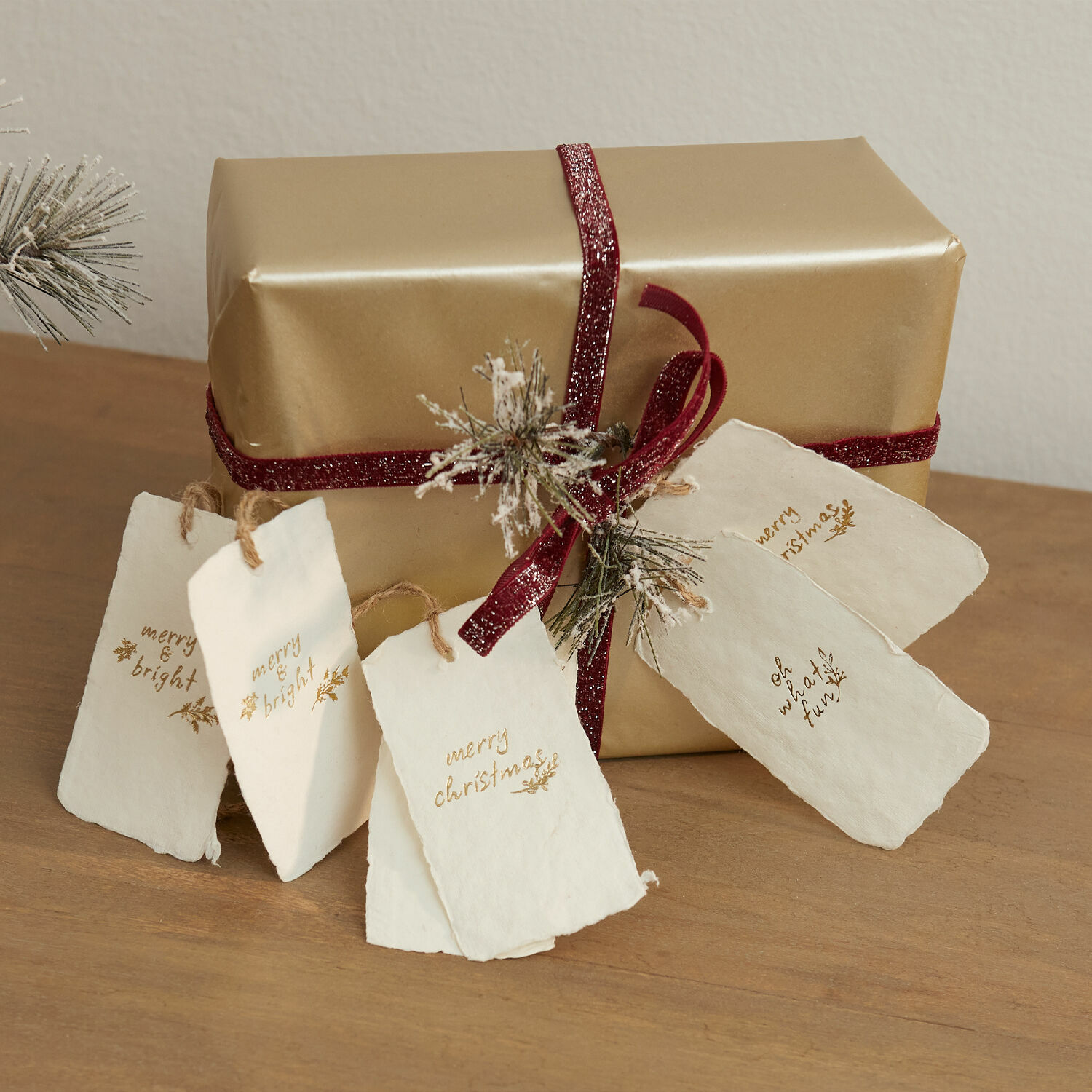 Anhänger 6er Set Aamira Goldverpacktes Geschenk mit bordeauxrotem Band und weißen Papieranhängern auf Holztisch