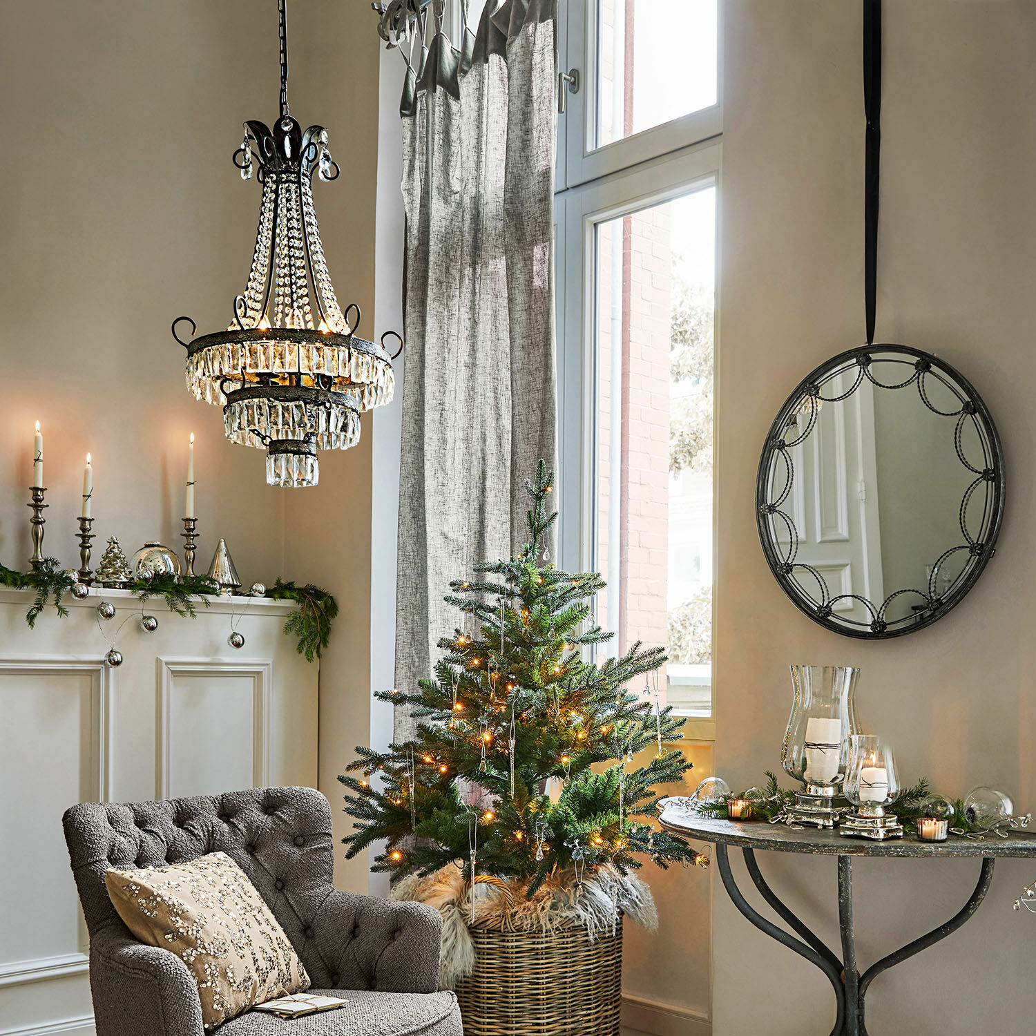 Gemütliches Wohnzimmer mit grauem Polstersessel, Weihnachtsbaum im Korb und Kristall-Kronleuchter