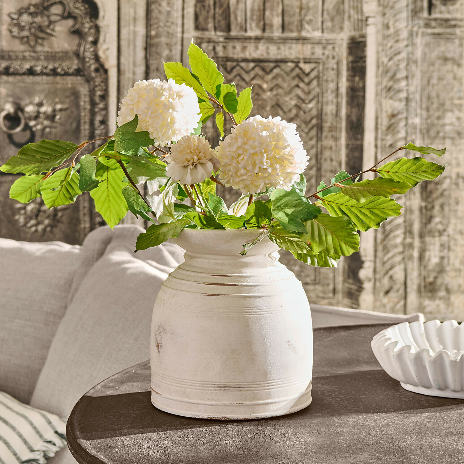 Vase Niverana Weiße Keramikvase mit grünen Blättern und cremefarbenen Blüten auf dunklem Vintage-Tisch