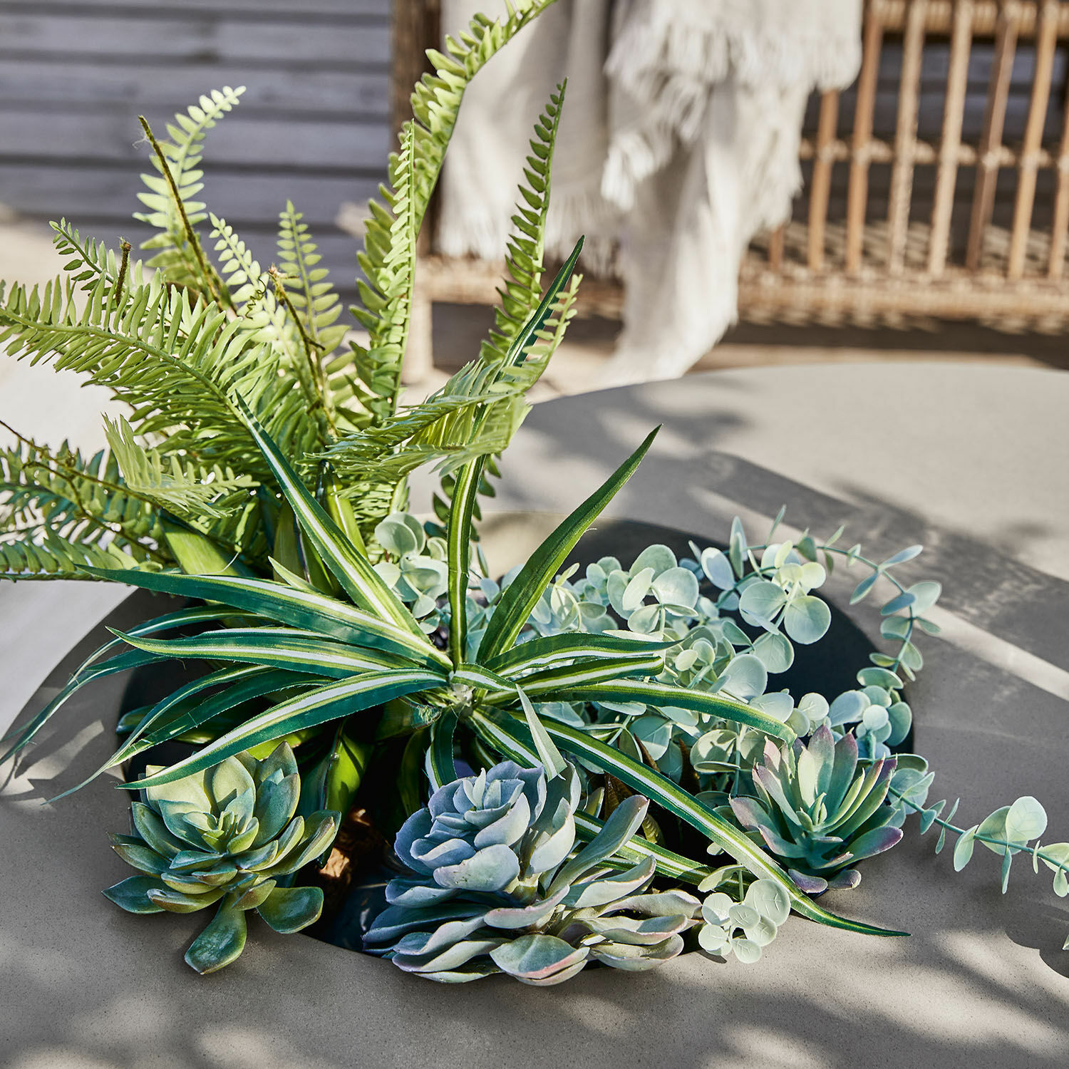 Arrangement aus künstlichen Sukkulenten und Farnen in Grüntönen auf grauem Gartentisch